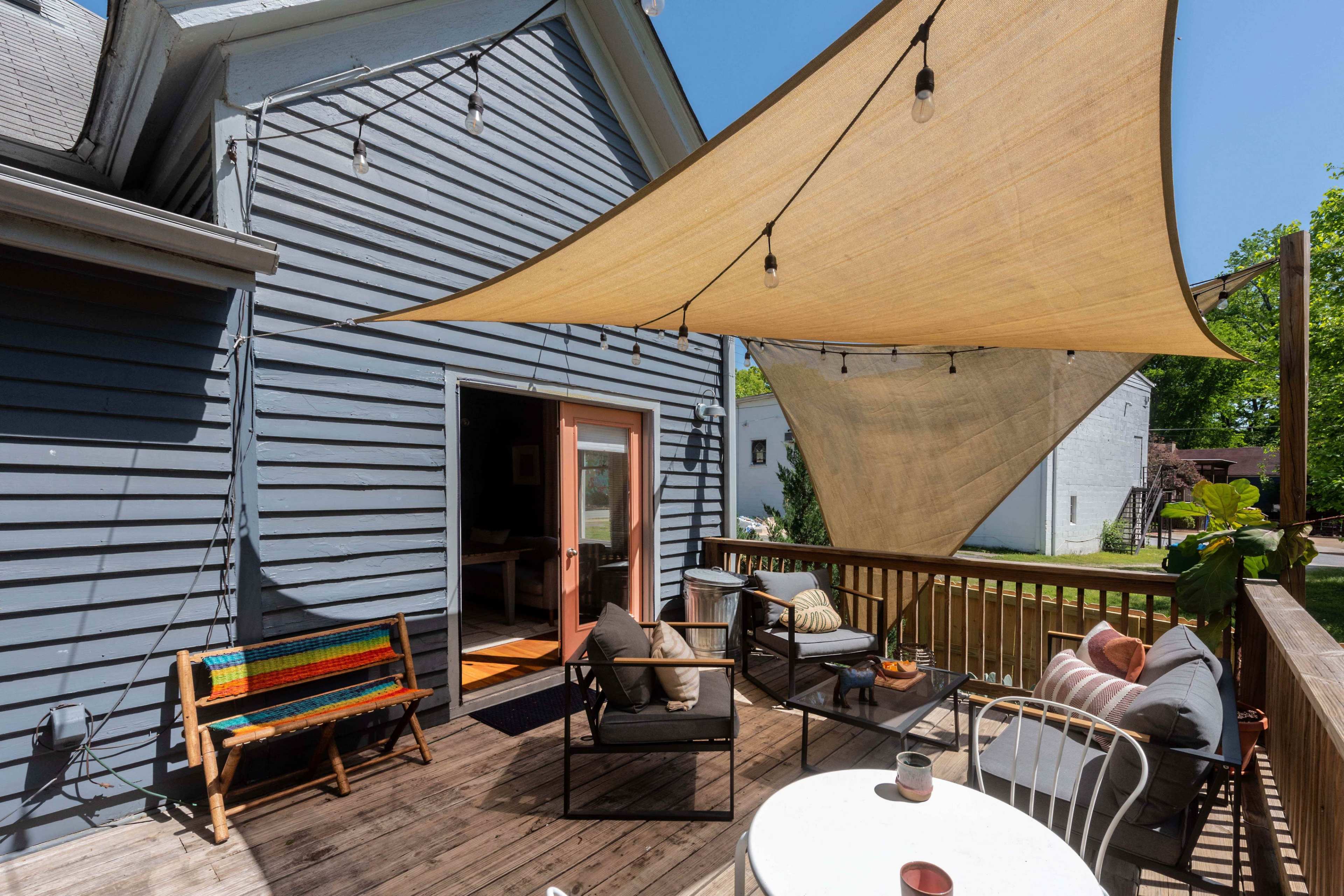 A wooden deck features a seating area with cushions, a small table, and a large shade sail overhead, creating a comfortable outdoor space.