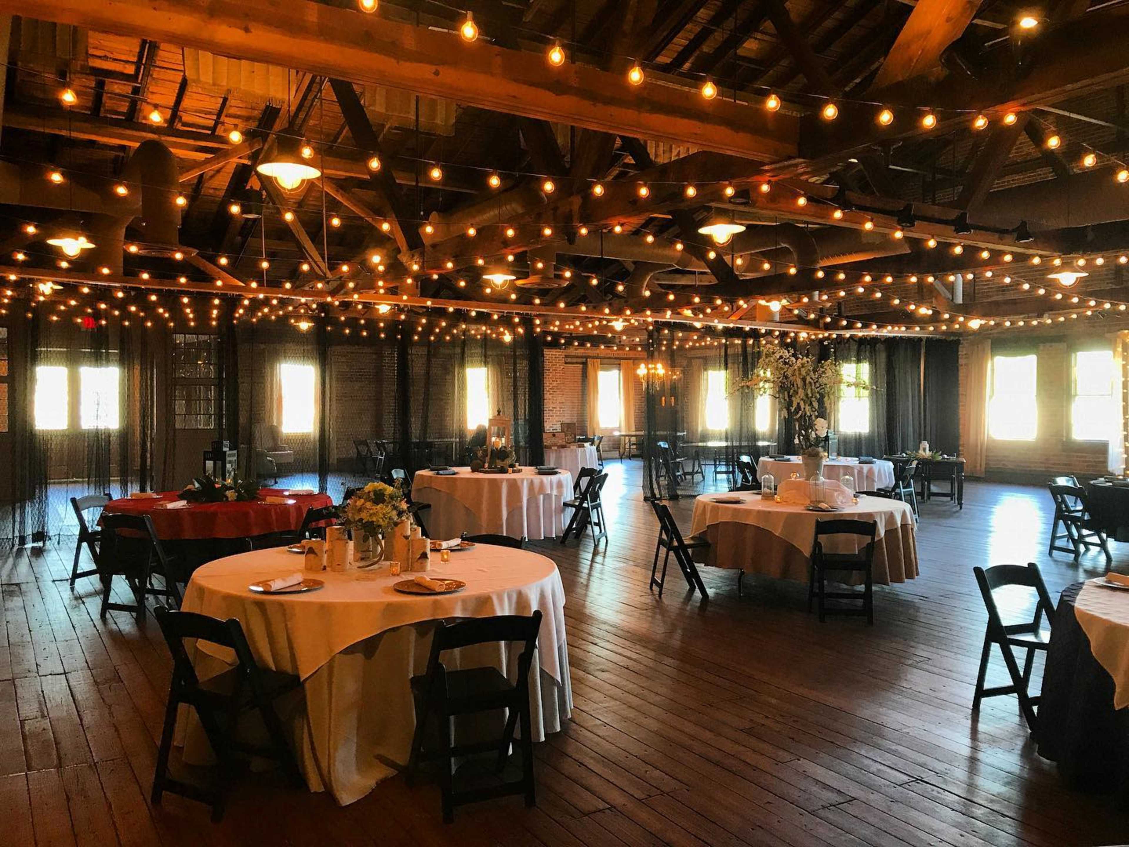 A spacious event hall with round tables set for dining, adorned with string lights overhead and large windows allowing natural light to enter.