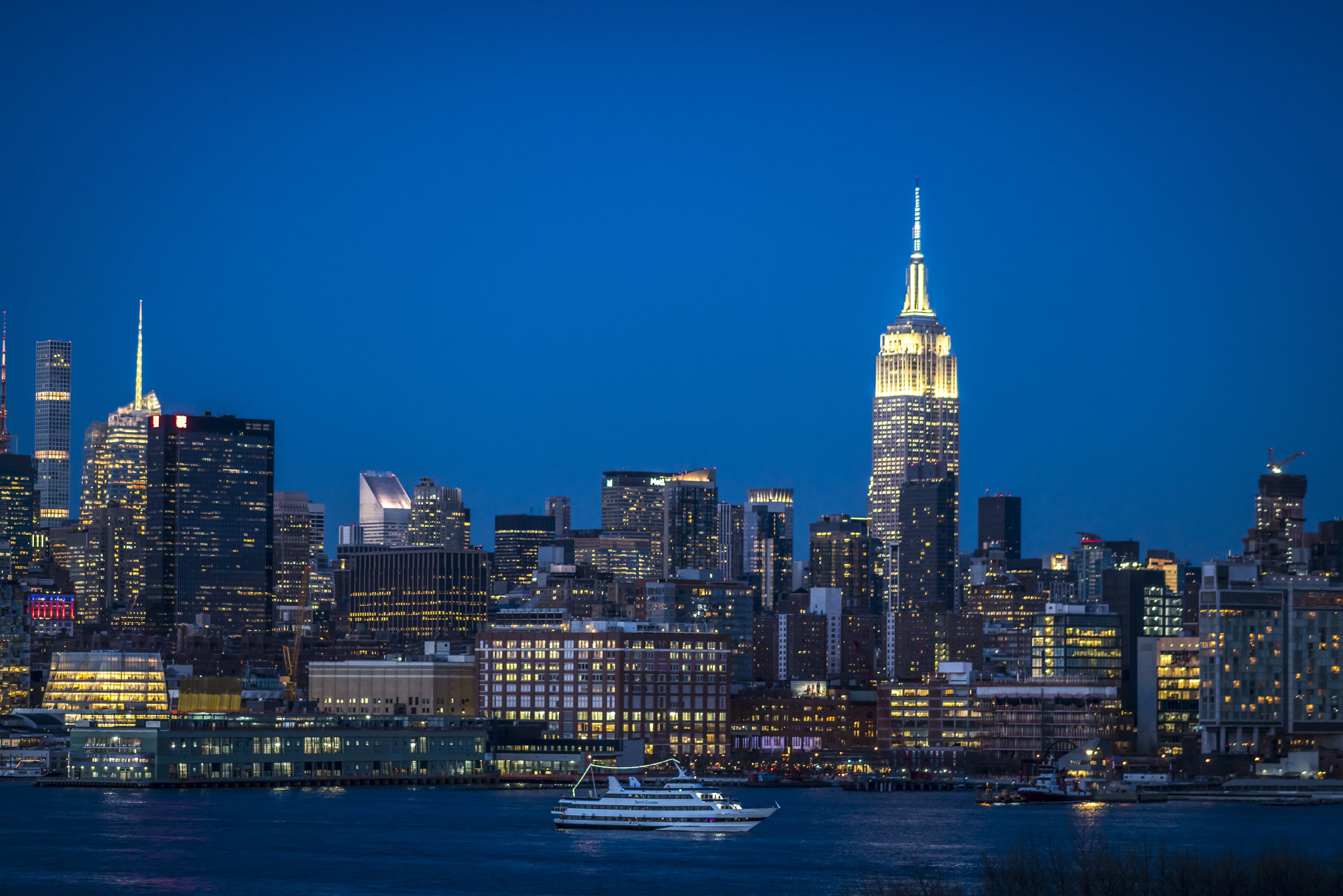 The Empire State Building illuminated against a twilight skyline of New York City, with a boat on the water in the foreground.