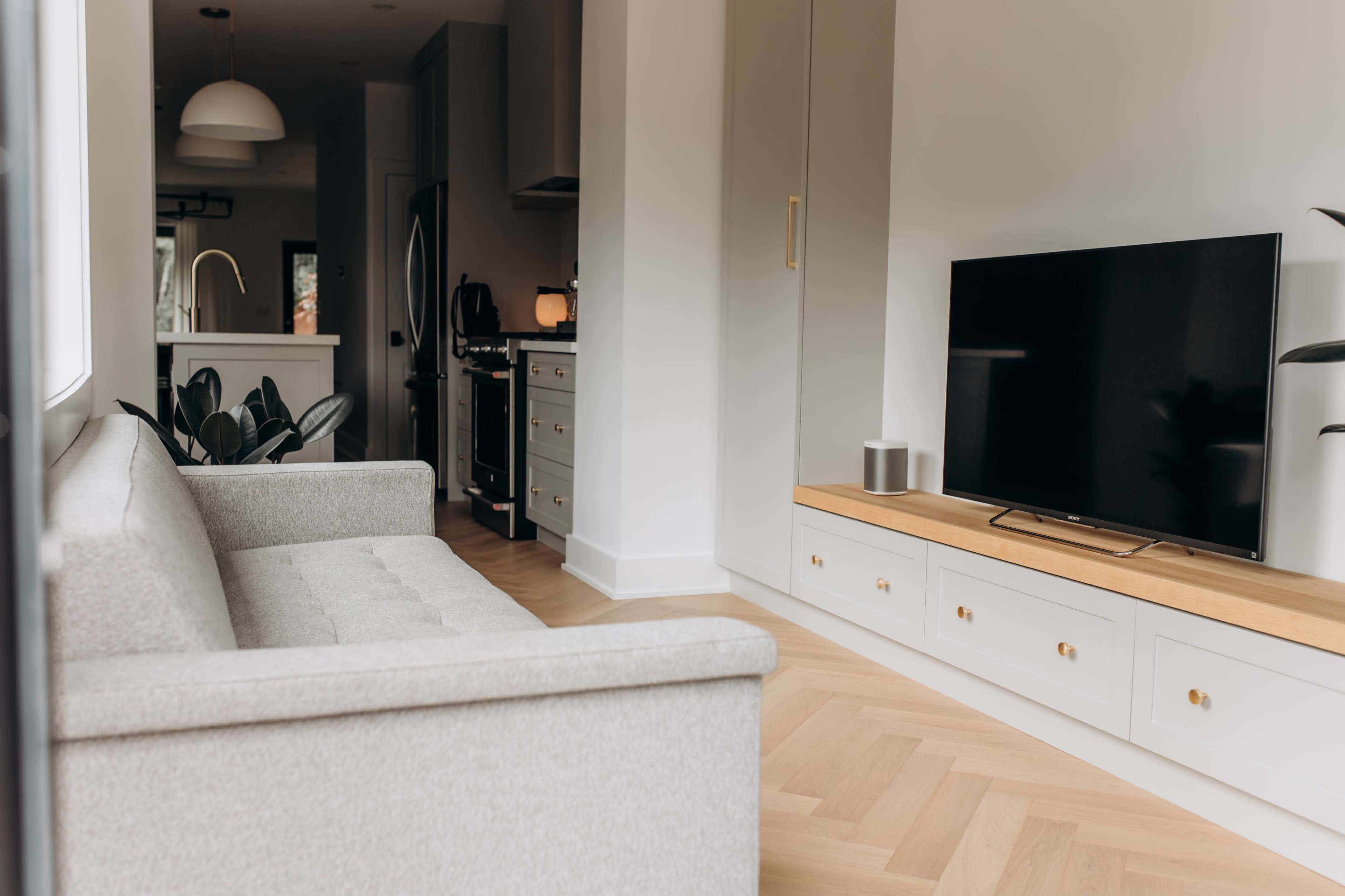 A modern living room with a light gray sofa facing a flat-screen TV on a wooden media console, adjacent to a kitchen area in the background.