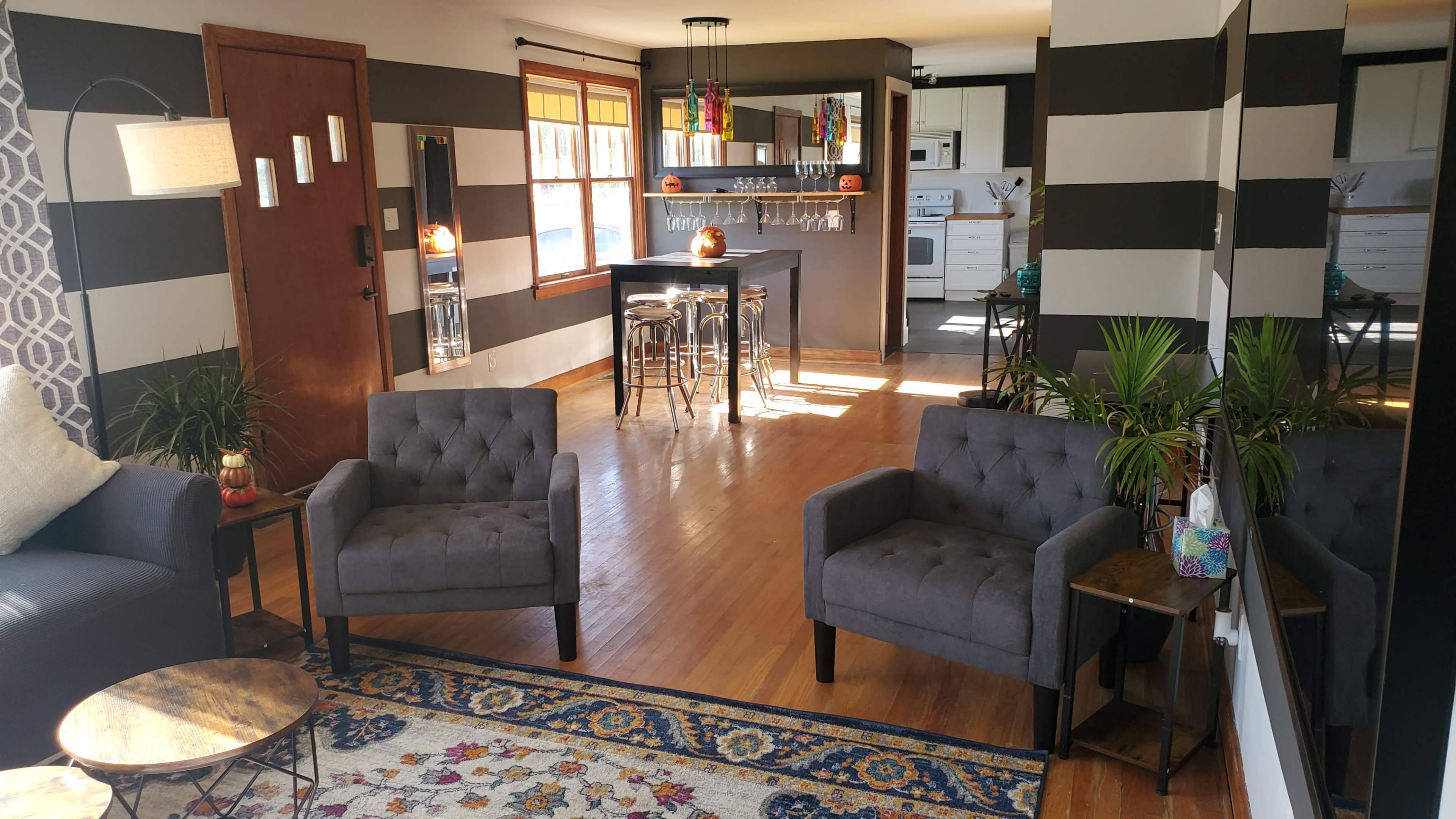 A bright living space with gray upholstered chairs, a wooden coffee table, a small bar area, and a kitchen visible in the background.