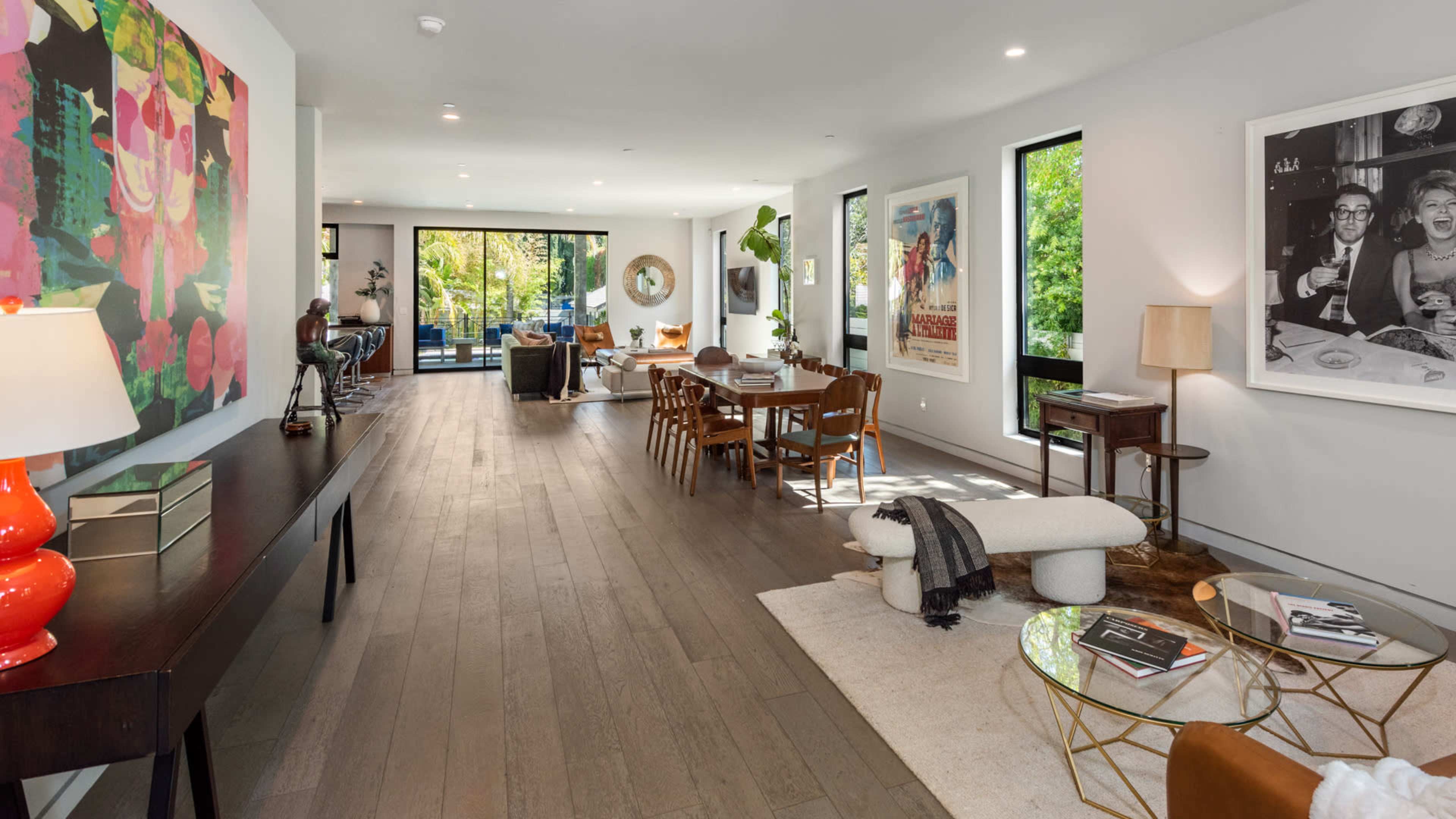The image shows a modern, open-concept living space featuring a dining area, a sitting area with a coffee table, large windows, and various decorative artworks on the walls.