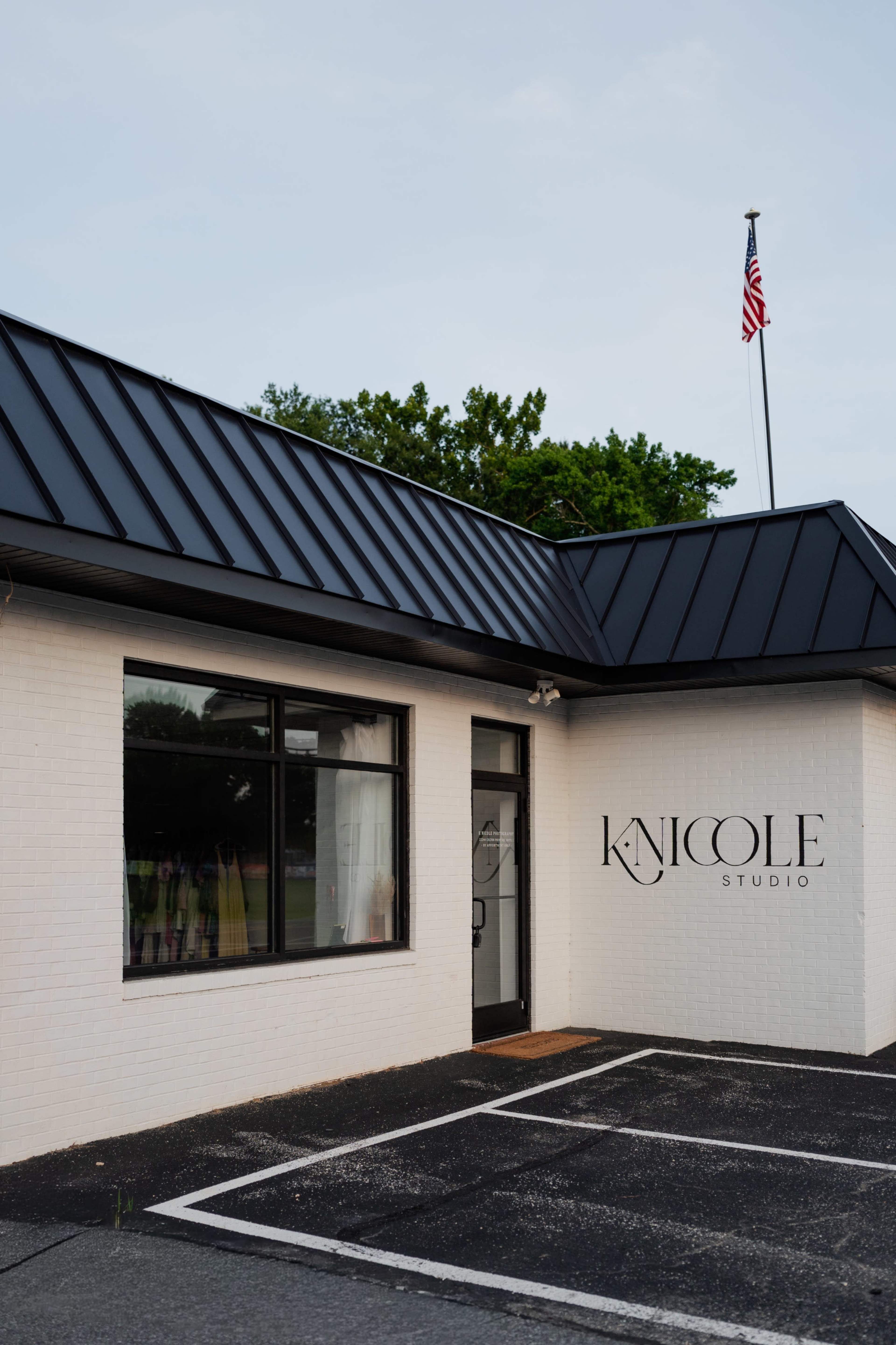 The image shows a white brick building with a black roof, featuring a sign that reads "NICOLE STUDIO" and a parking area in front.