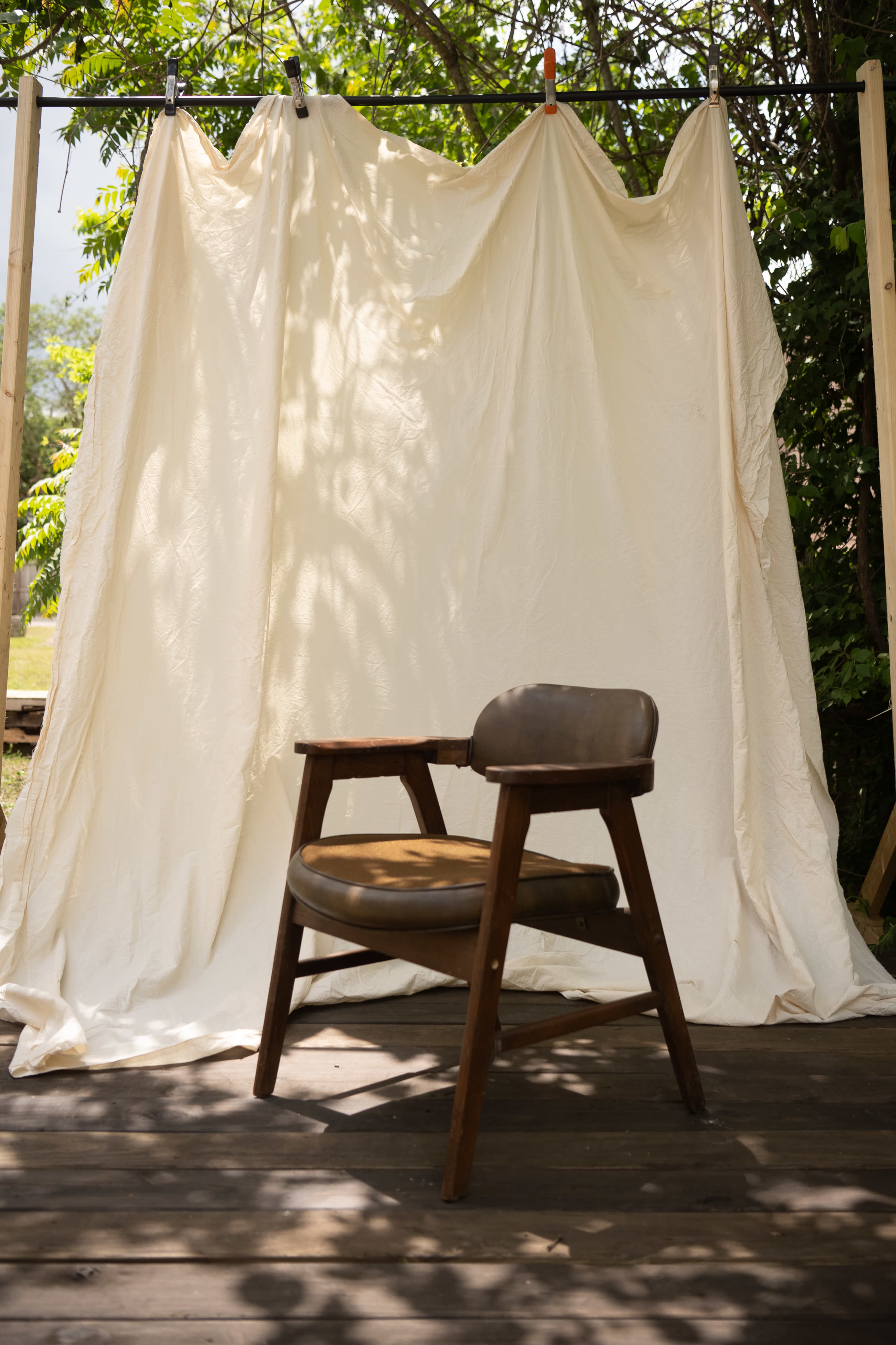A wooden chair with a brown seat is positioned in front of a white draped backdrop amongst greenery.