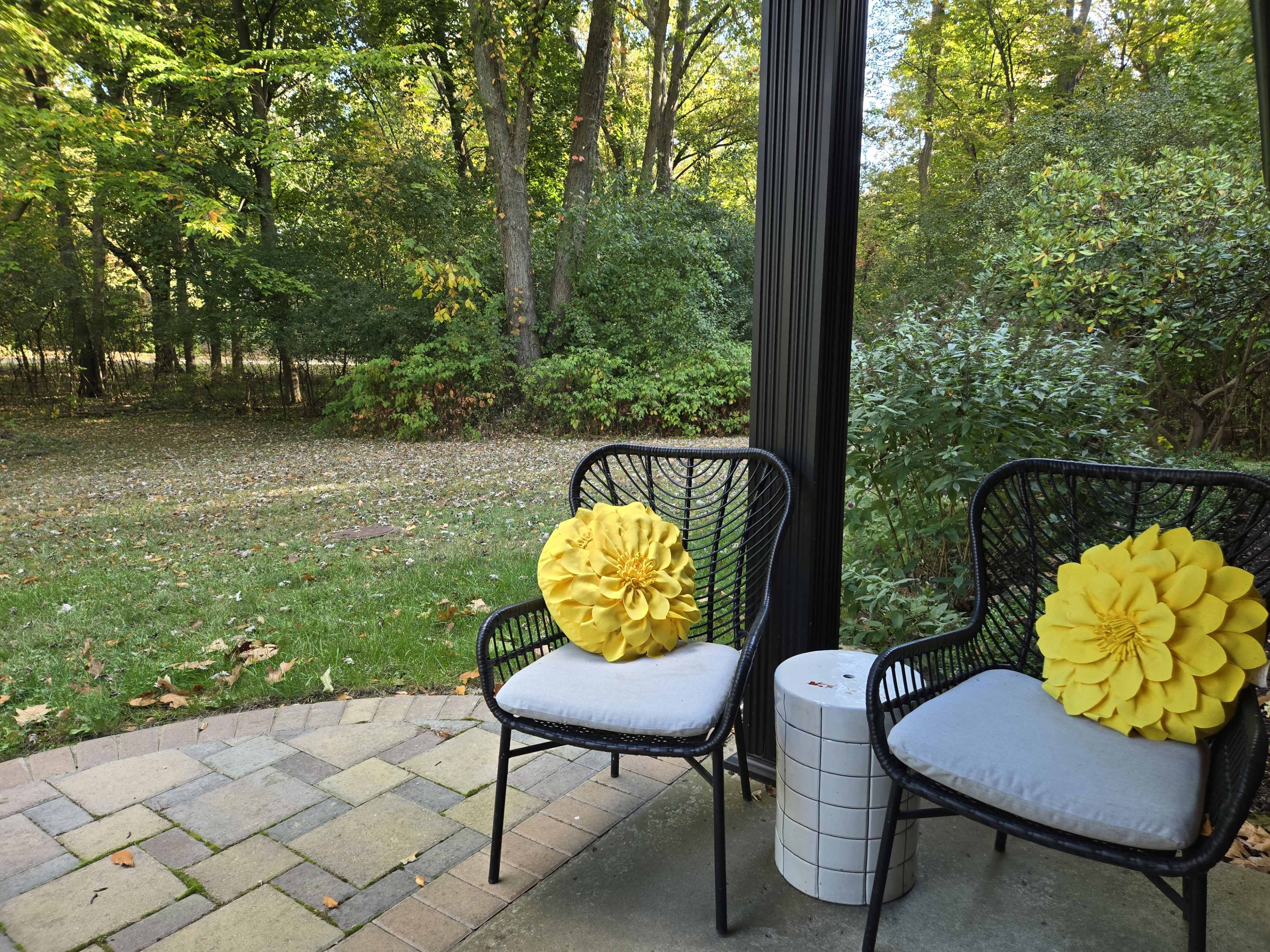 Two black wicker chairs with gray cushions and yellow decorative pillows are placed on a paved patio overlooking a grassy area with trees in the background.