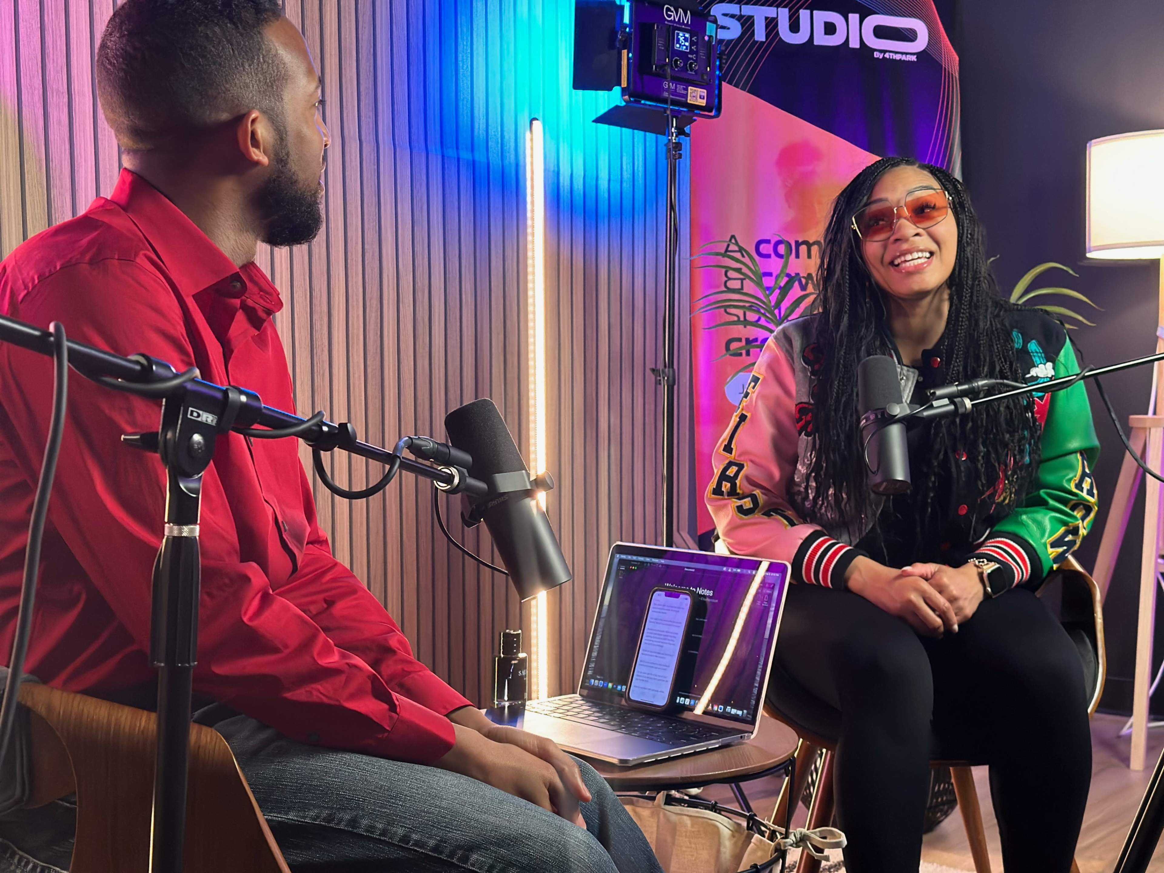 A man and woman are sitting in front of microphones, engaged in conversation in a well-lit studio setting.