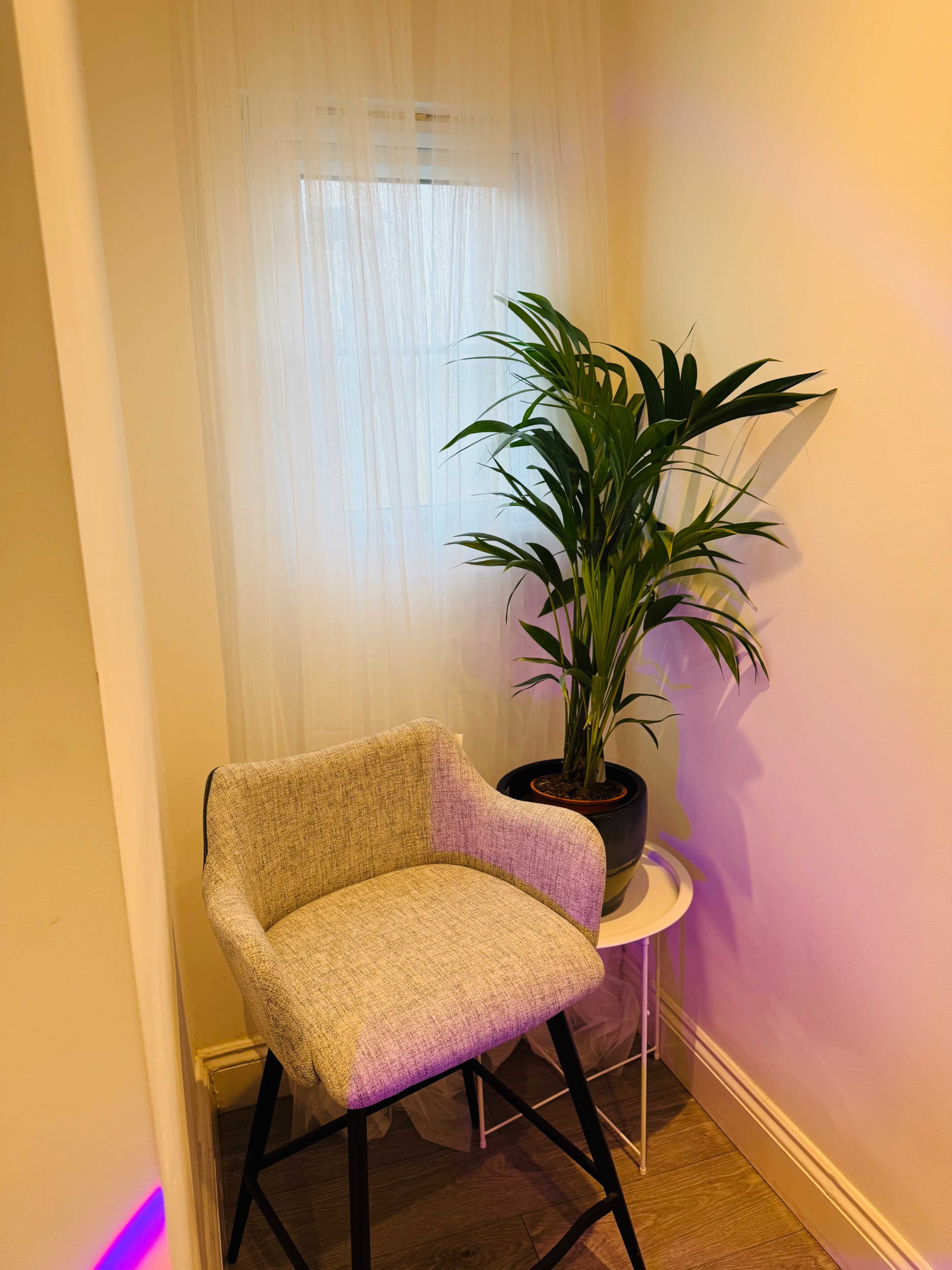 A gray upholstered bar stool is positioned next to a potted plant on a small white table in a well-lit corner of a room.