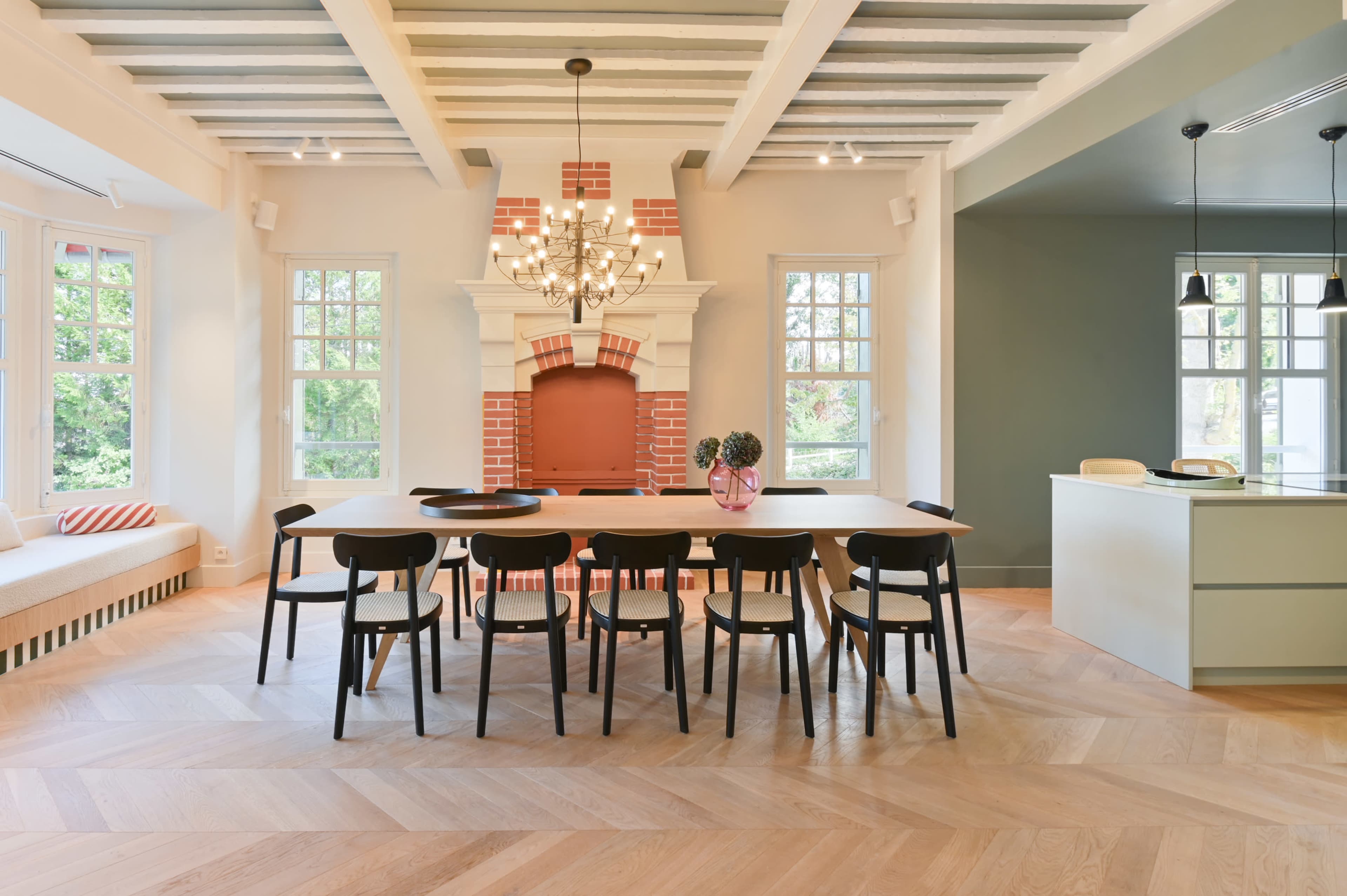 A dining area features a long table surrounded by black chairs, with a large fireplace and large windows letting in natural light.