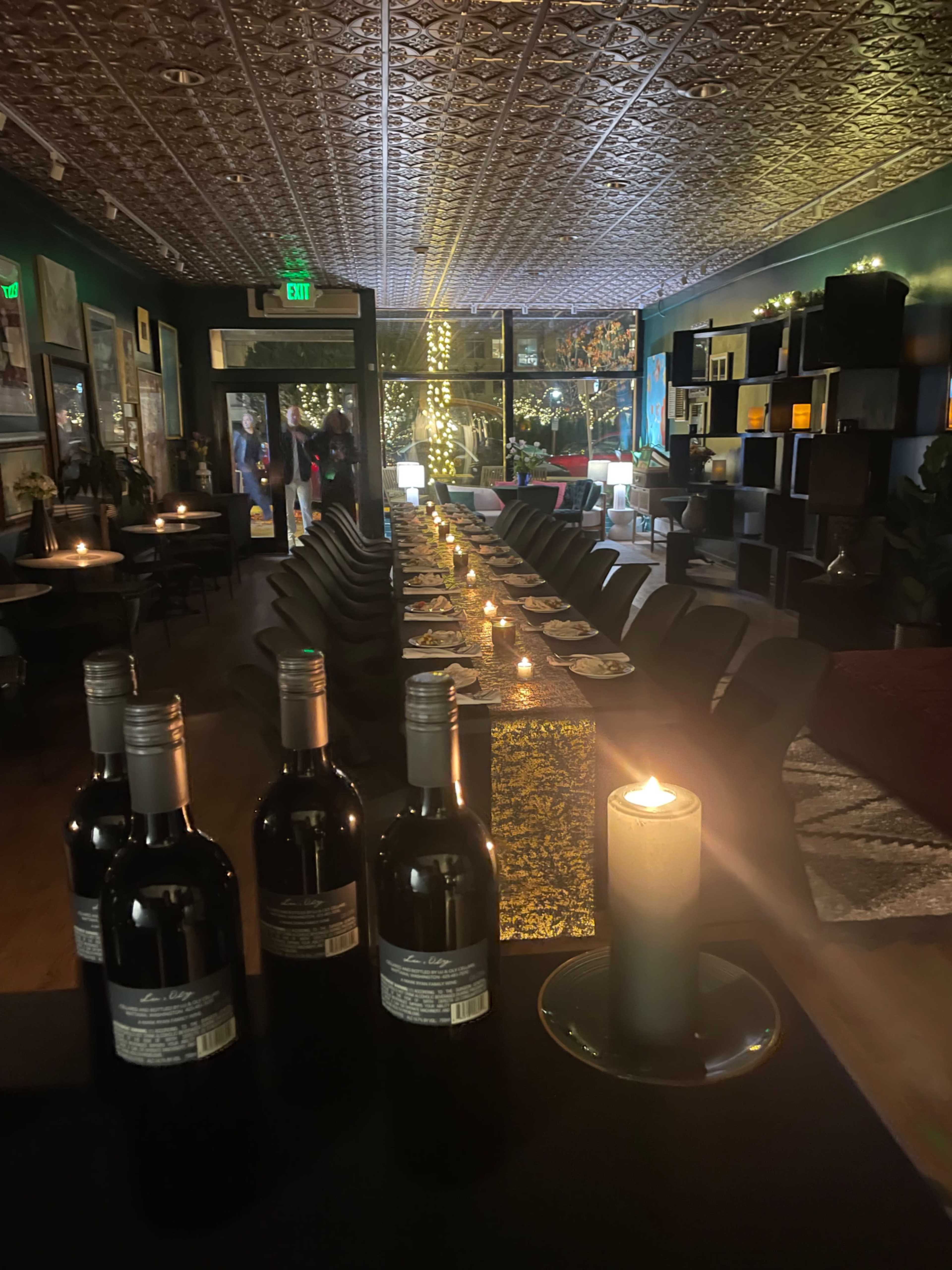 A long table is set with candles and wine bottles in a dimly lit restaurant, featuring dark walls and decorative accents.