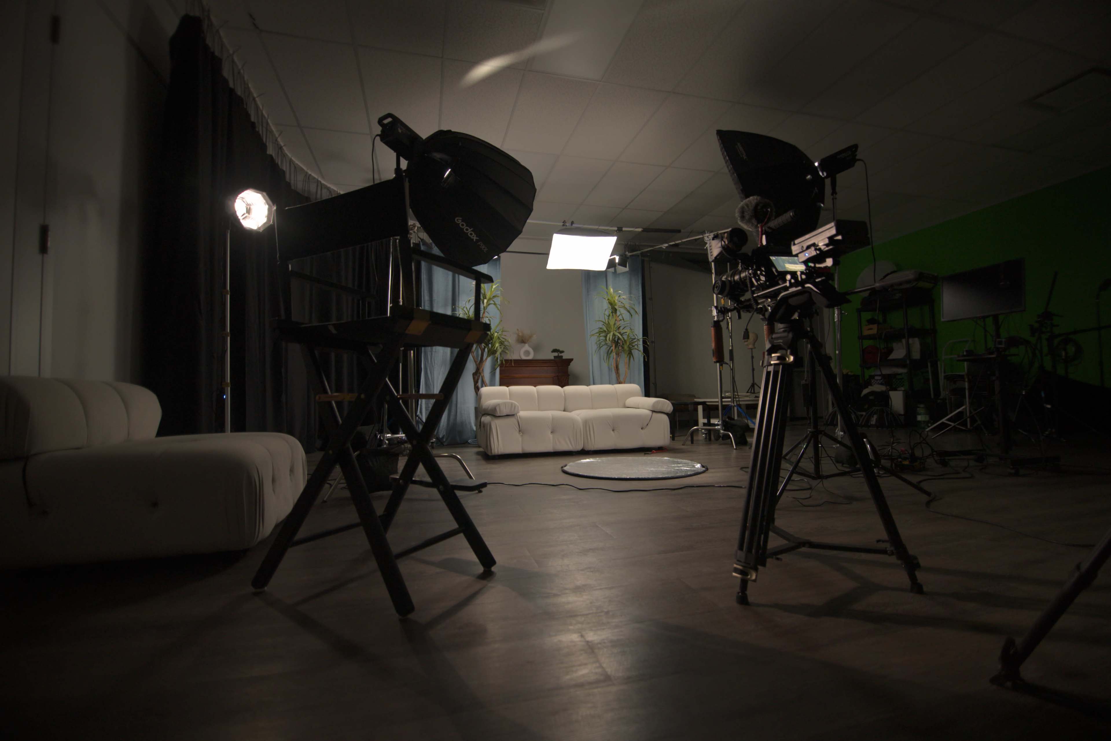 The image shows a darkened studio setup with lighting equipment, a camera on a tripod, and a white sofa in the background.
