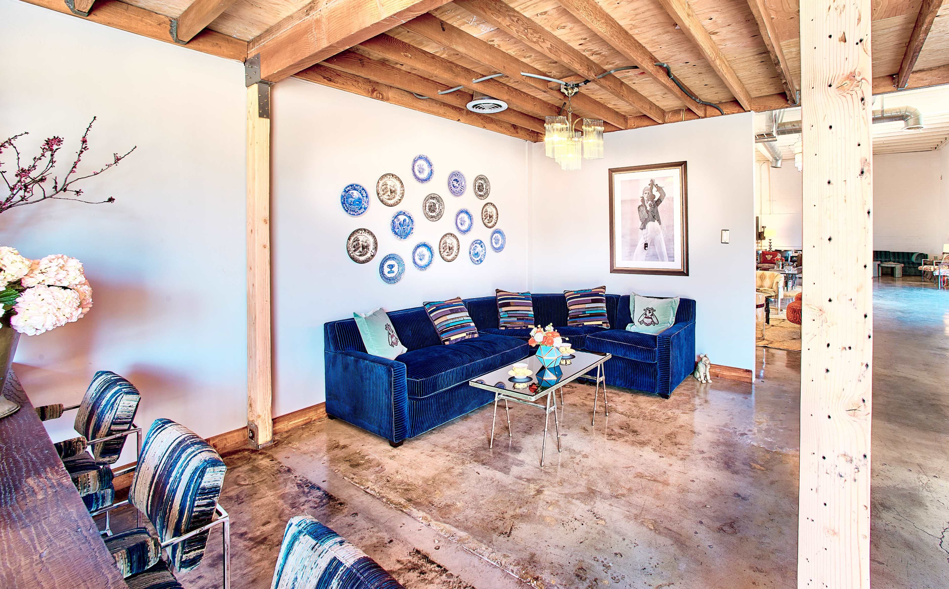 A living room features a blue sectional sofa adorned with striped cushions, a glass coffee table, and decorative wall plates, all beneath exposed wooden beams and natural light.