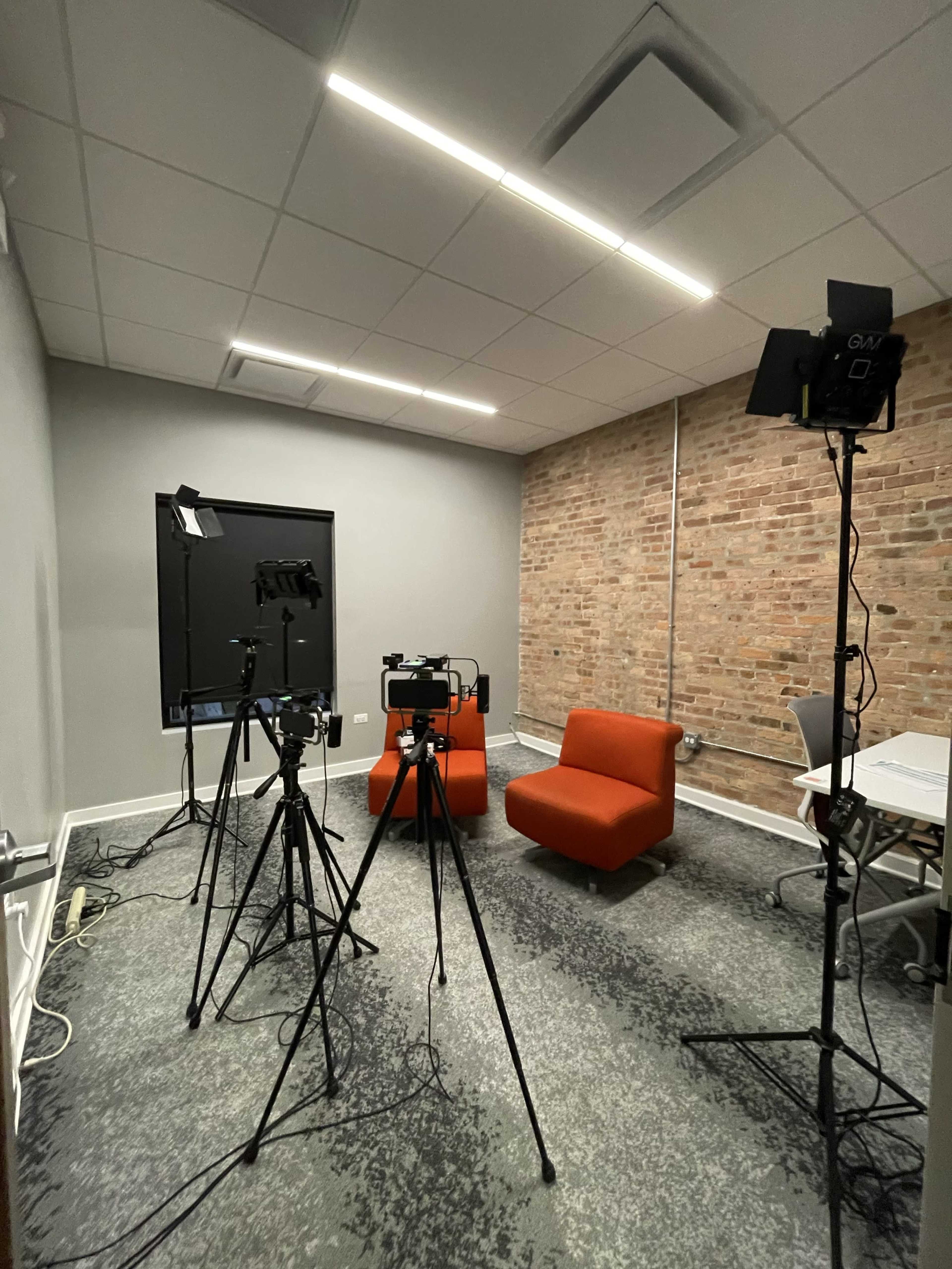 A small recording studio features two orange chairs, three tripods with cameras, and a backdrop against a brick wall.