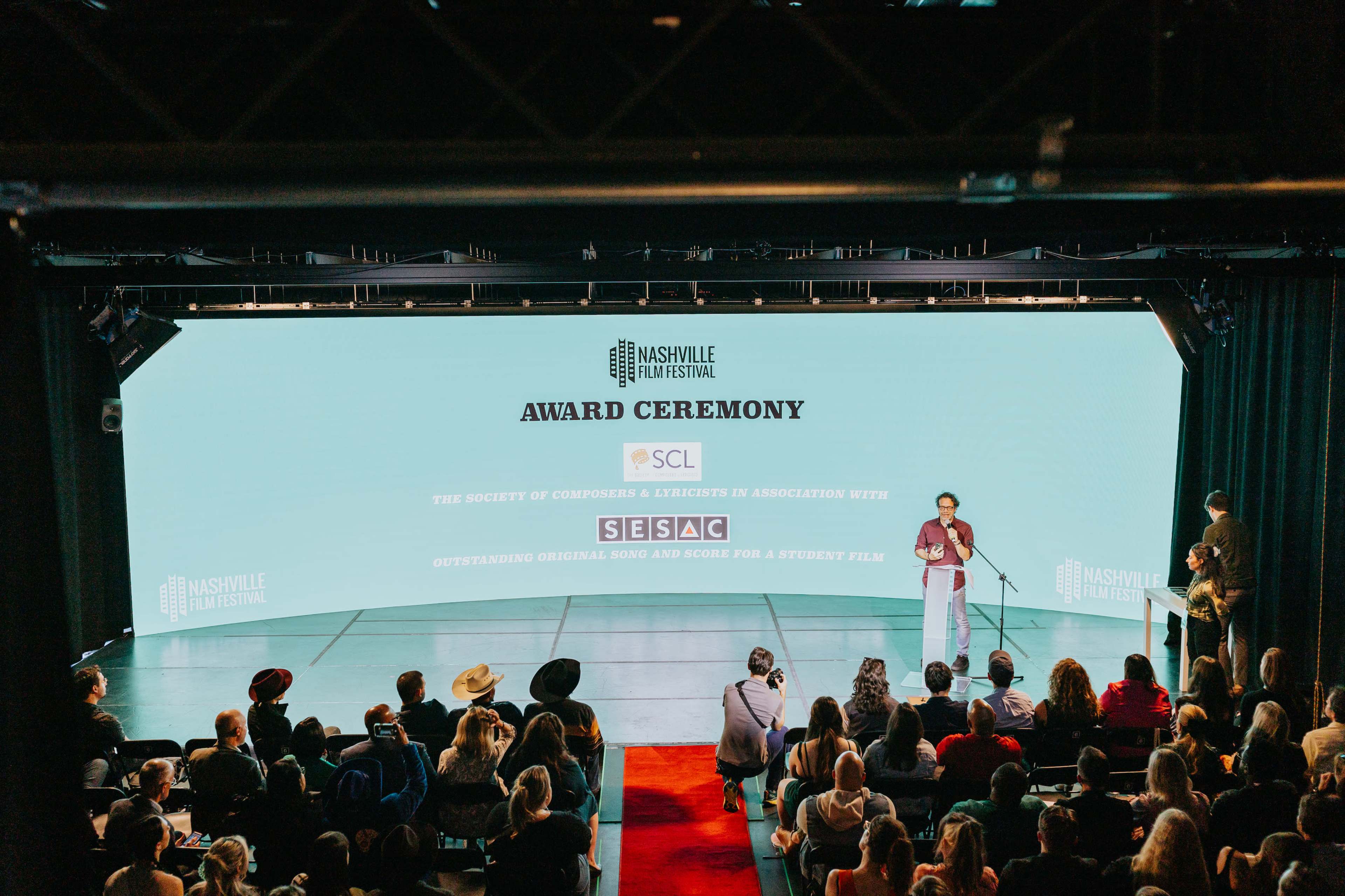 A speaker delivers a presentation at an award ceremony with an audience seated in front of a large screen displaying event details.