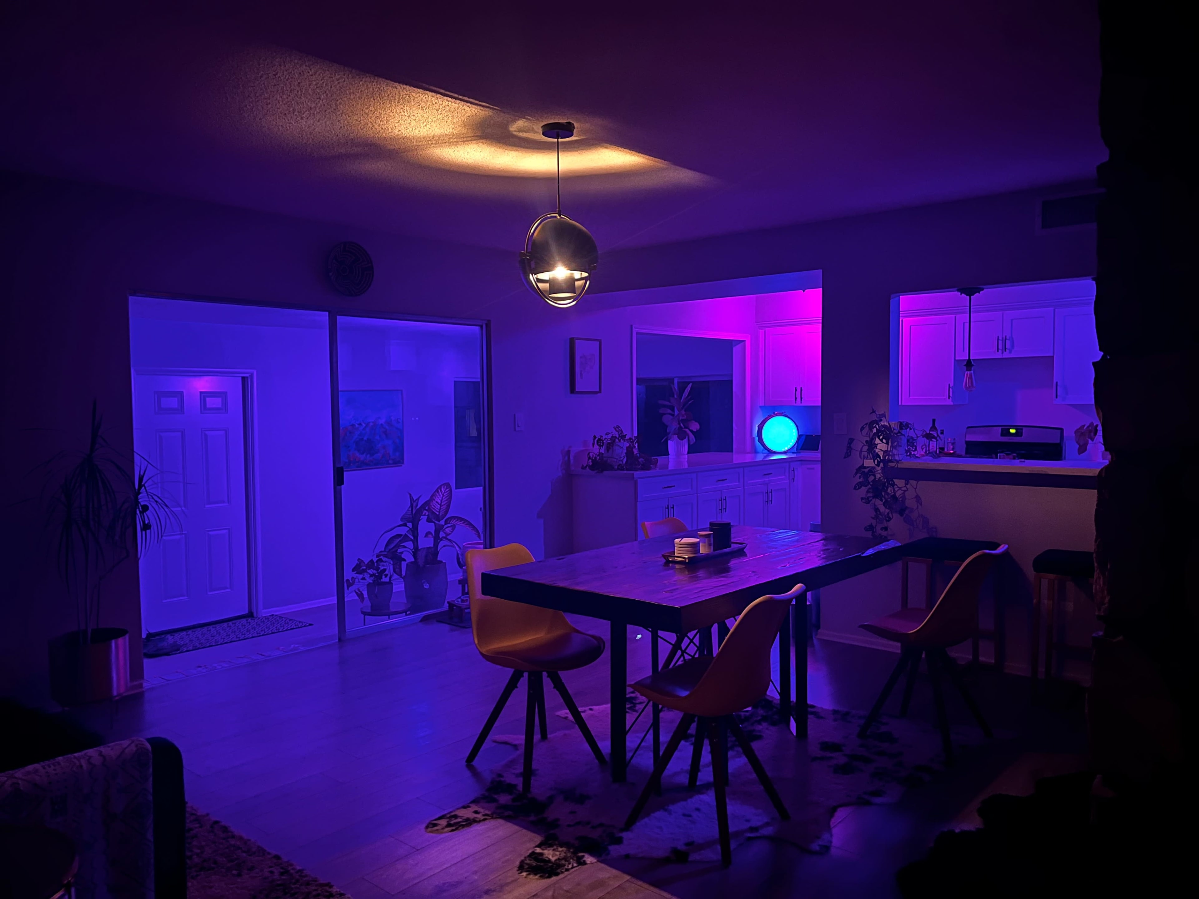 A dimly lit dining area with a large table surrounded by colorful purple and blue lighting, highlighting the kitchen and entryway in the background.