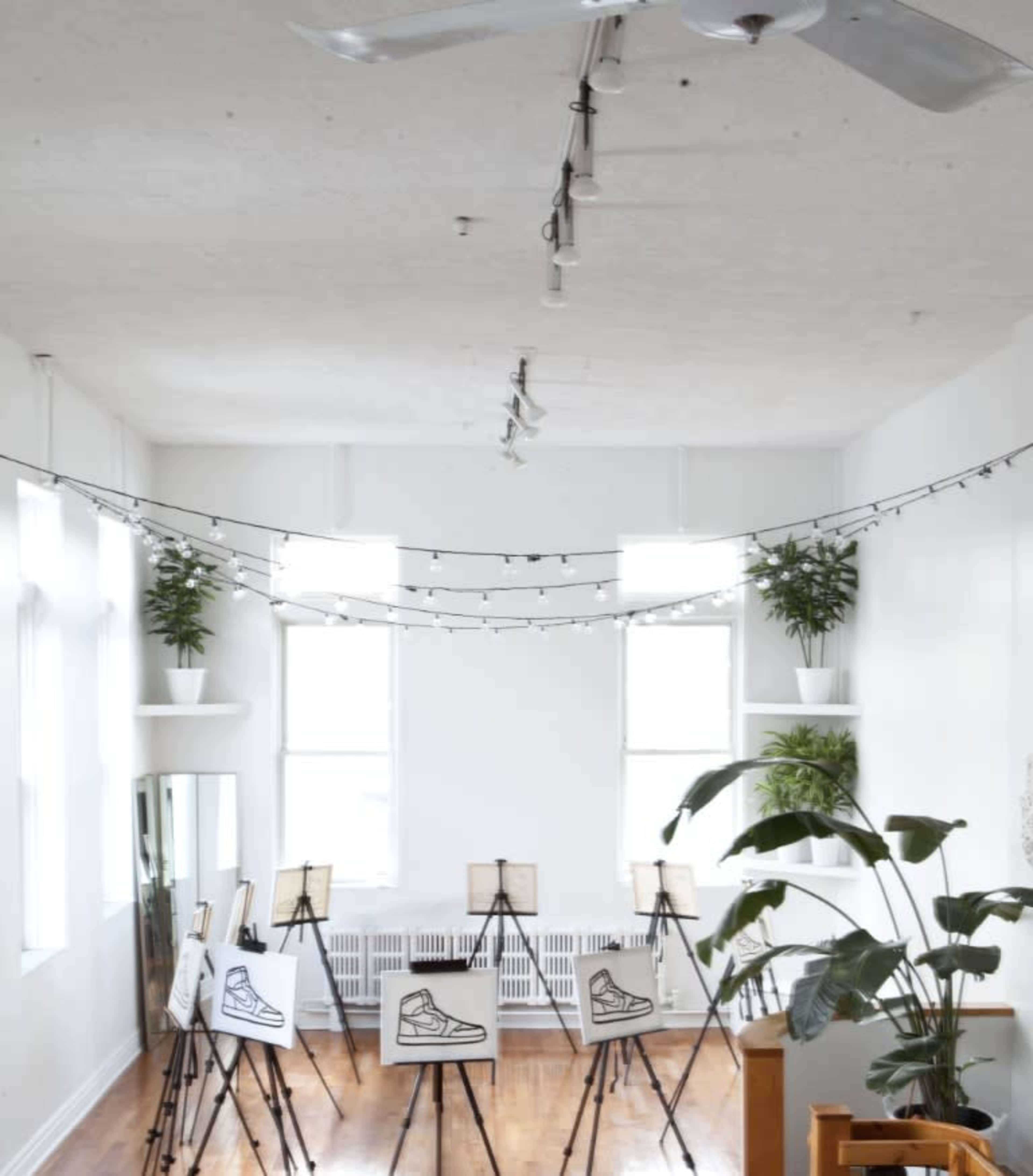 A spacious room features multiple easels displaying drawings of shoes, with potted plants and string lights accentuating the bright, airy space.