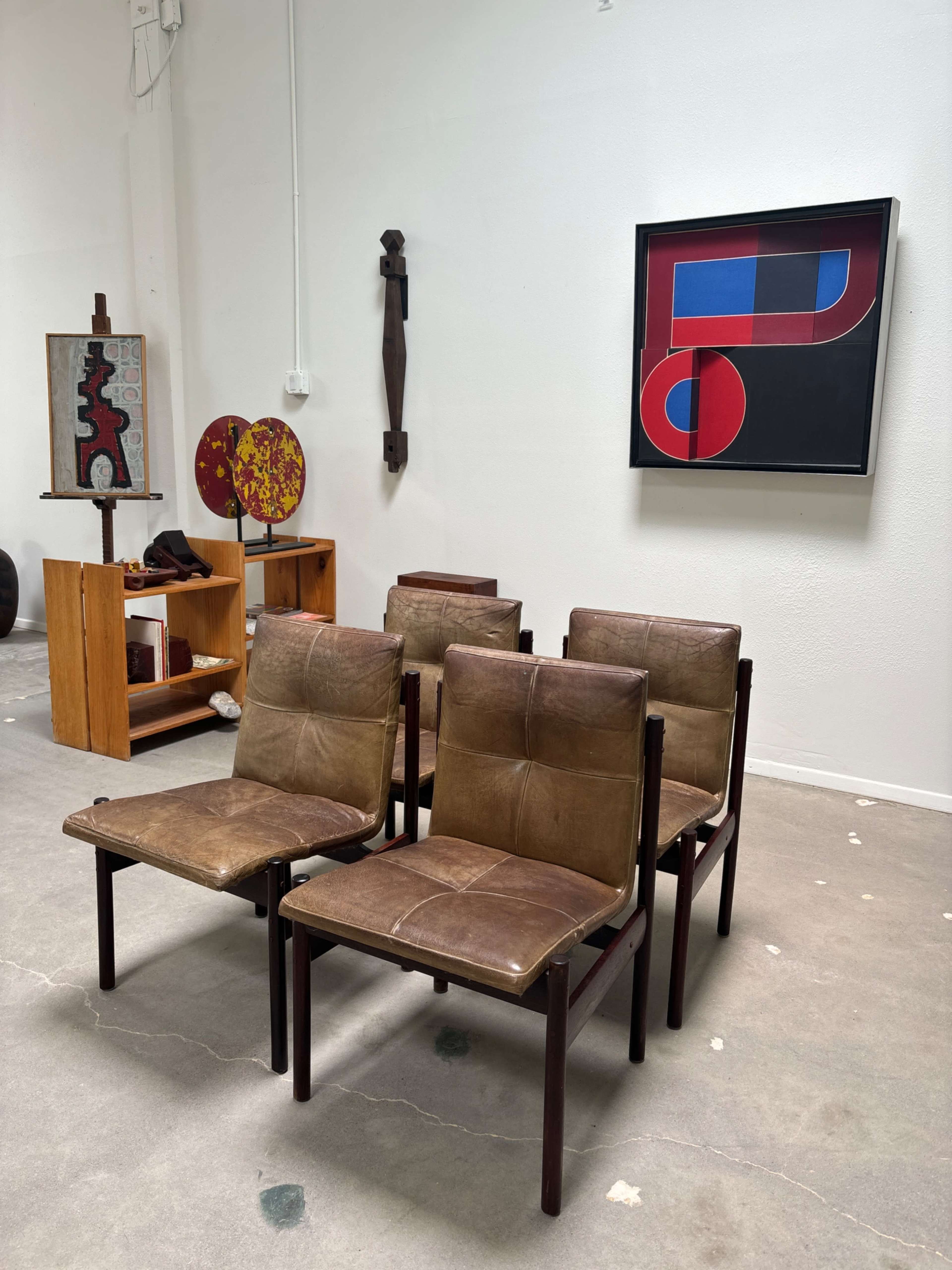 A set of four brown, leather chairs arranged in front of a white wall, alongside various artworks and furniture in an art gallery setting.