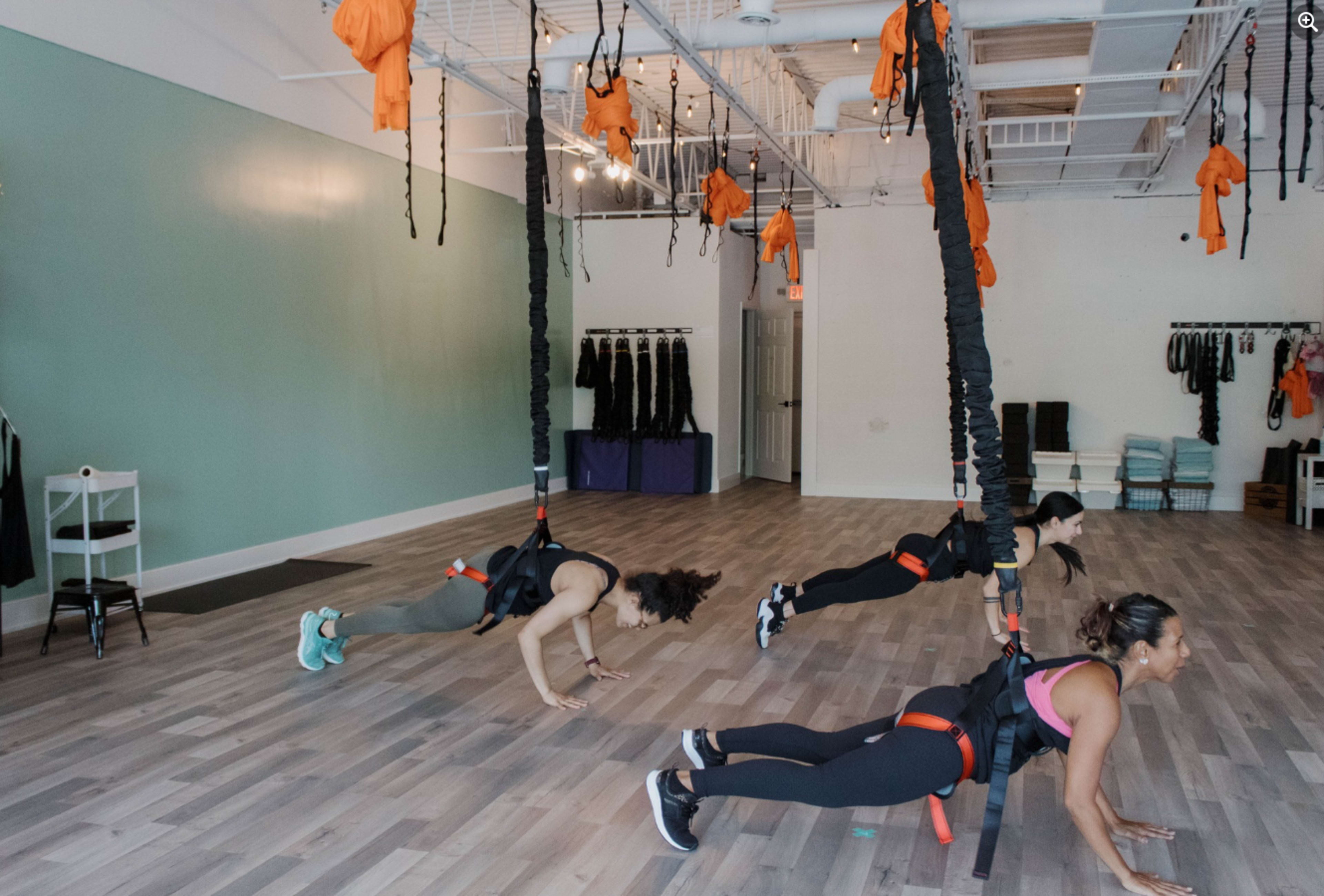 Three individuals are performing an exercise routine using suspension straps in a fitness studio with a light green wall and wooden flooring.