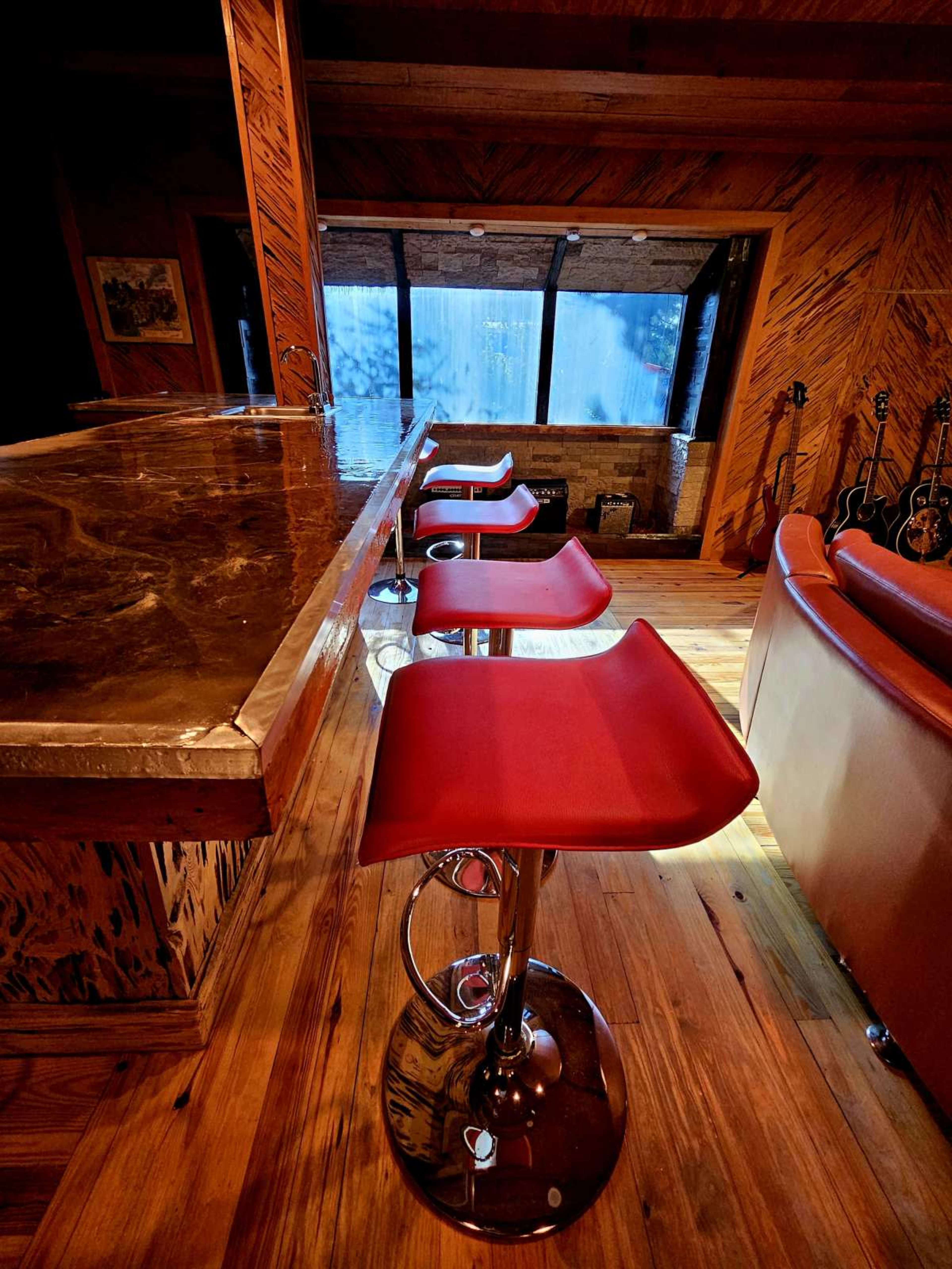 The image shows four red bar stools positioned around a wooden countertop in a room with large windows displaying a view of water or an outdoor scene.