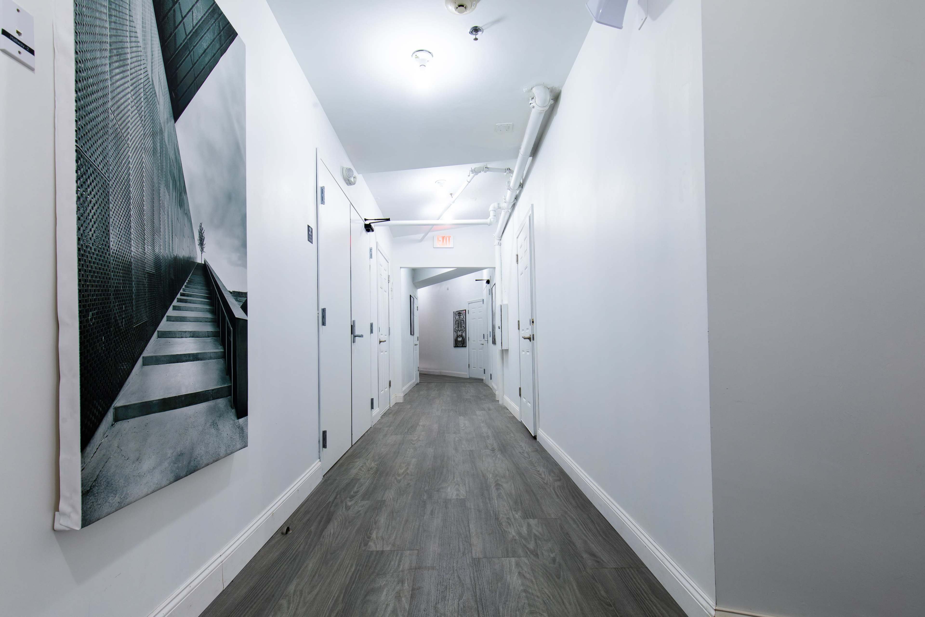 The image shows a long, narrow hallway with white walls and a textured floor, featuring doors on both sides and a large black-and-white photograph on the left wall.