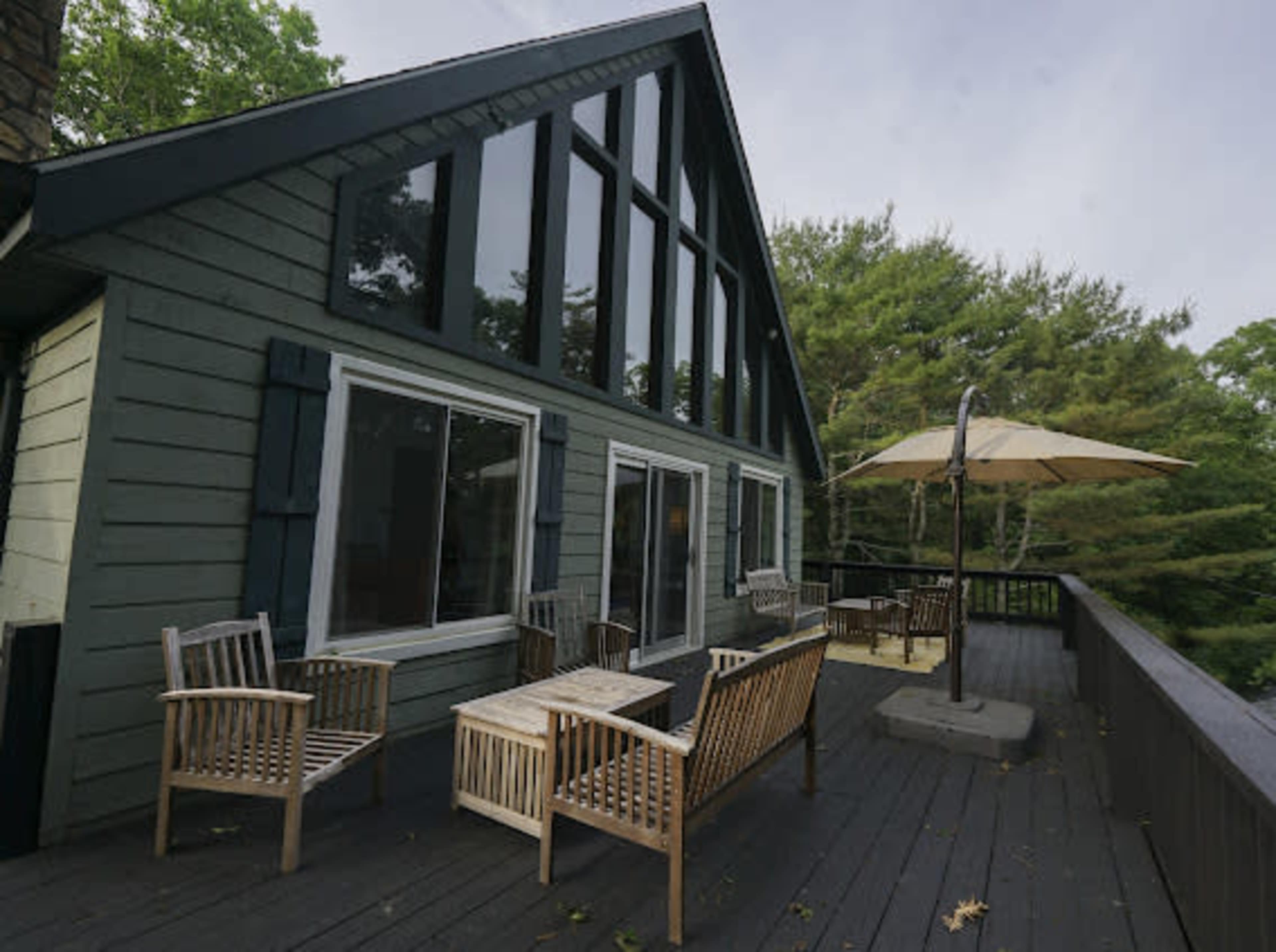 The image shows a green, A-frame house with large windows and a deck furnished with wooden chairs and a parasol.