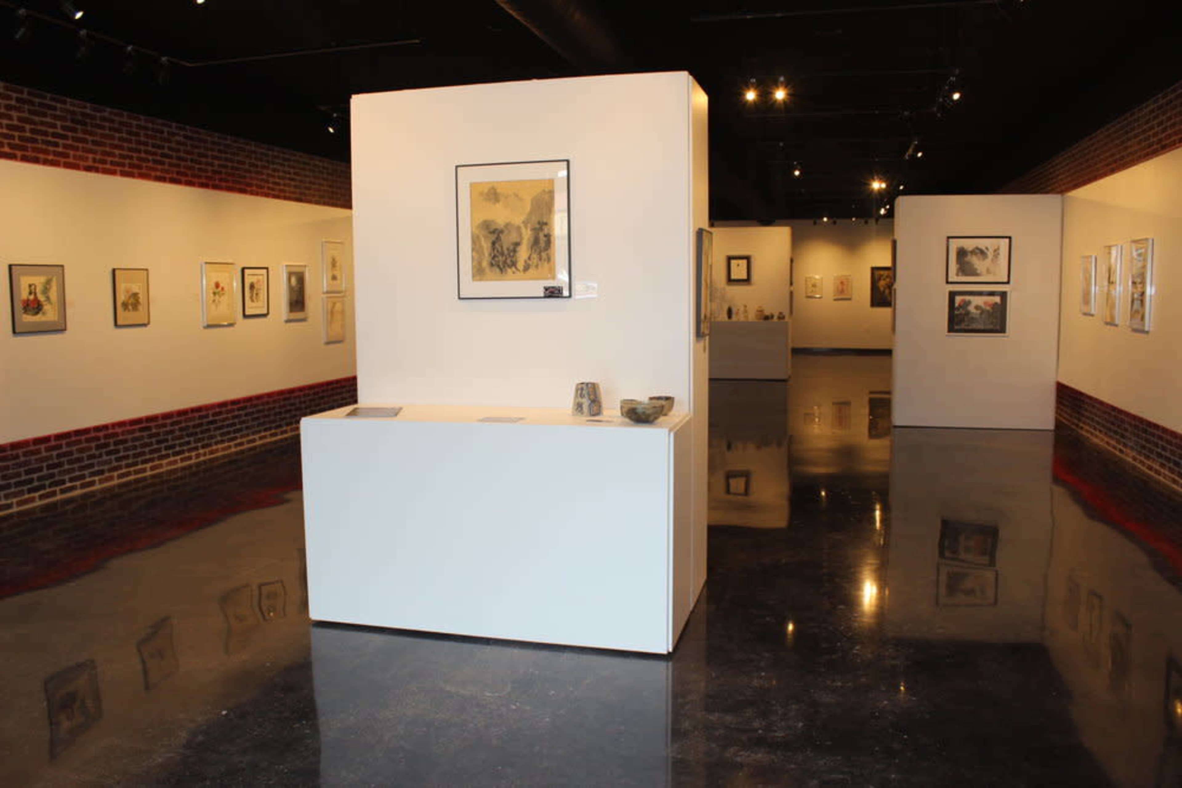 An art gallery with white walls displaying various framed artworks and a central pedestal holding additional pieces.