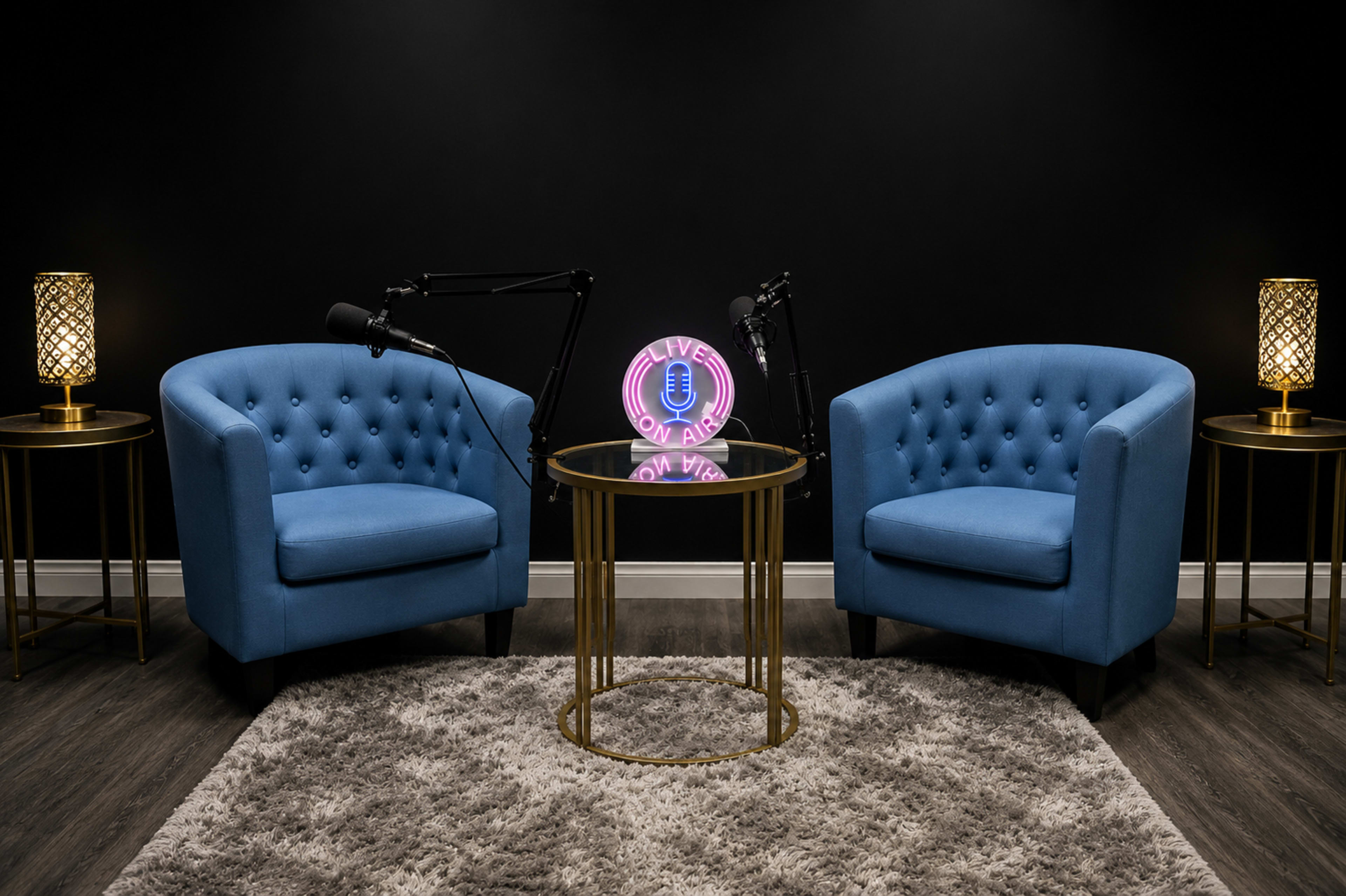 The image shows a modern podcast studio setup featuring two blue tufted chairs, a glass coffee table, and a neon microphone sign against a black wall, with two microphones mounted on stands.