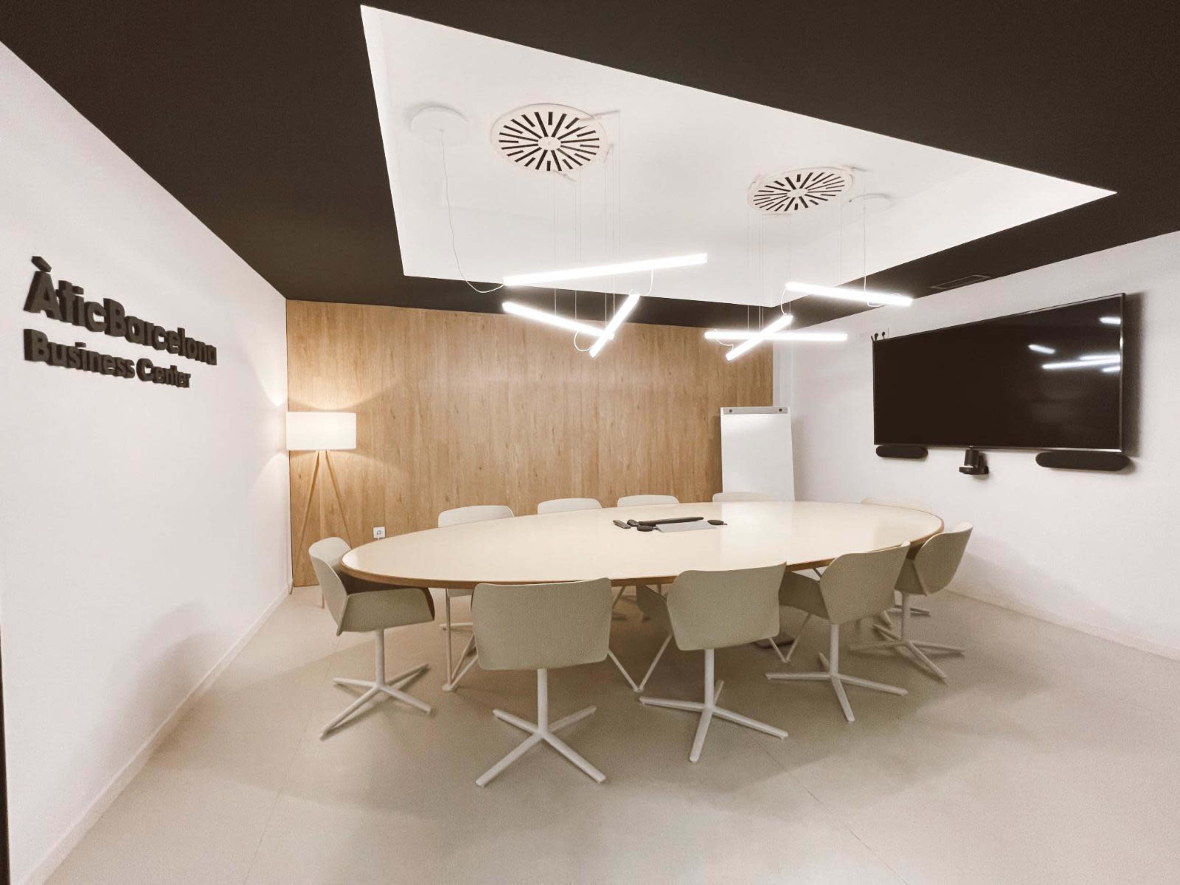 A modern conference room features a circular table surrounded by light-colored chairs, a wall-mounted television, and contemporary lighting.