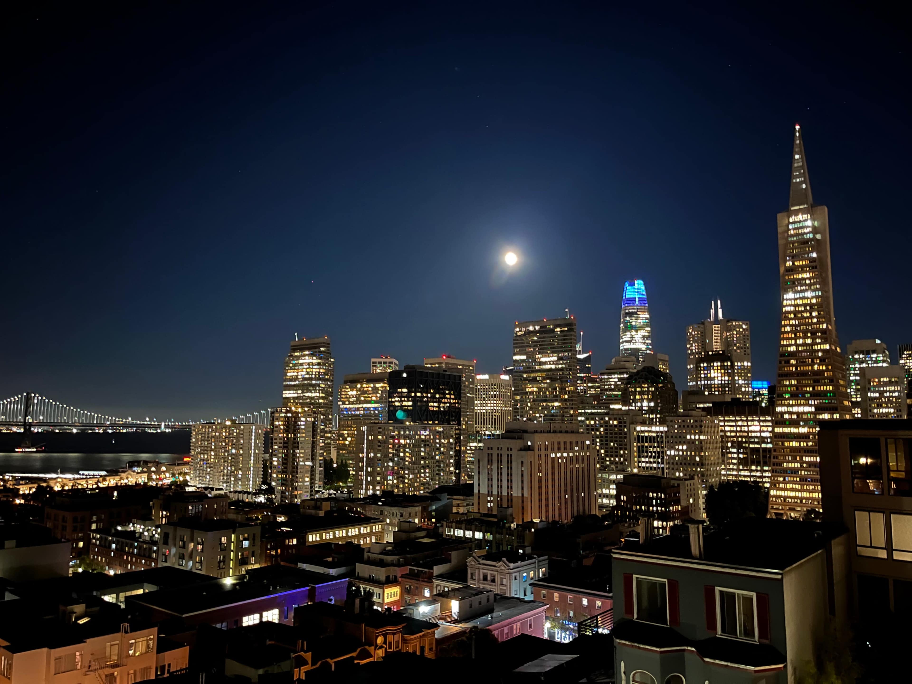 A nighttime view of San Francisco's skyline featuring illuminated buildings and the Bay Bridge in the background with a full moon overhead.