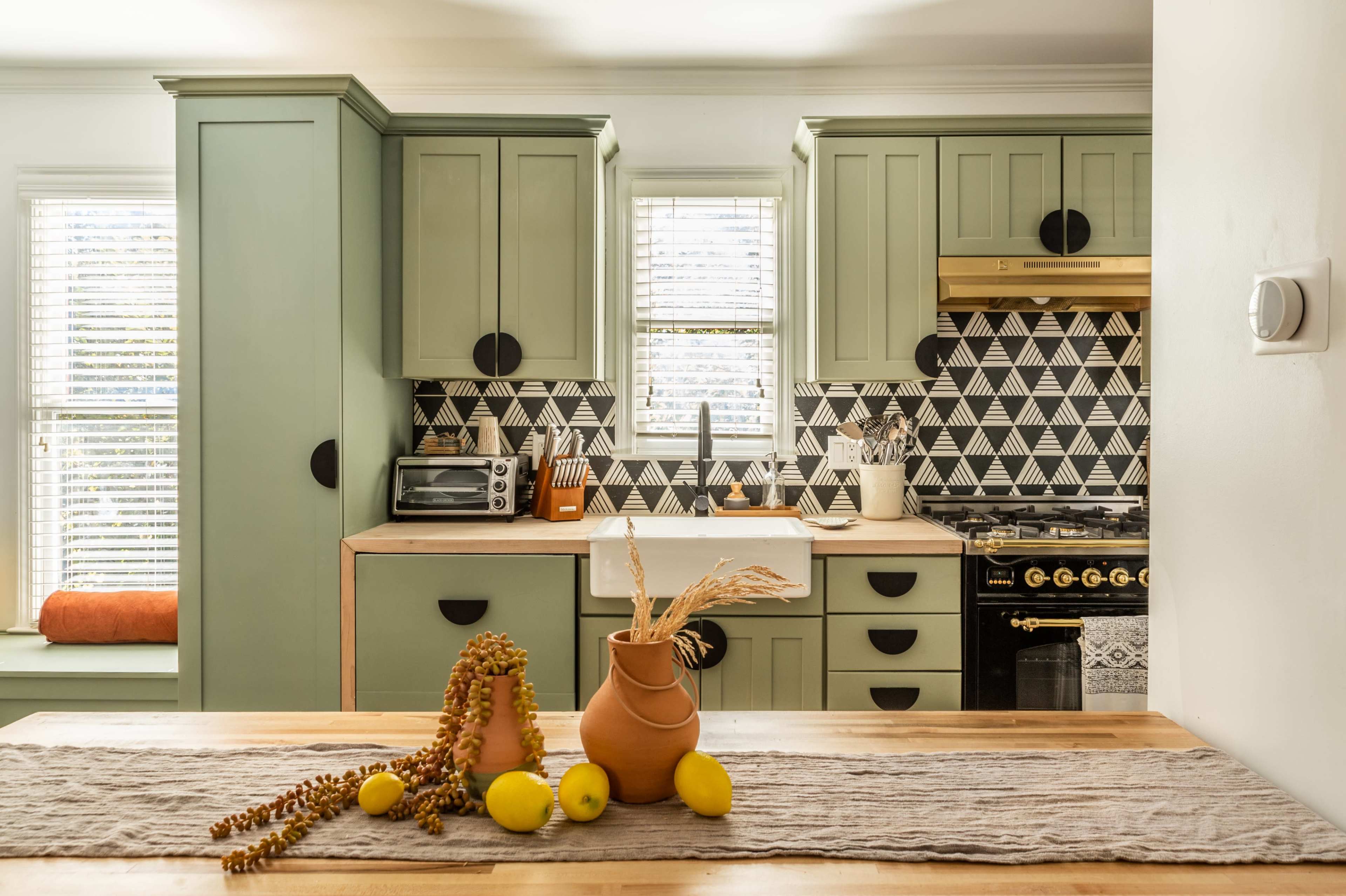 A modern kitchen features green cabinetry, a black stove, a white sink, an array of decorative items on the counter, and a patterned backsplash.