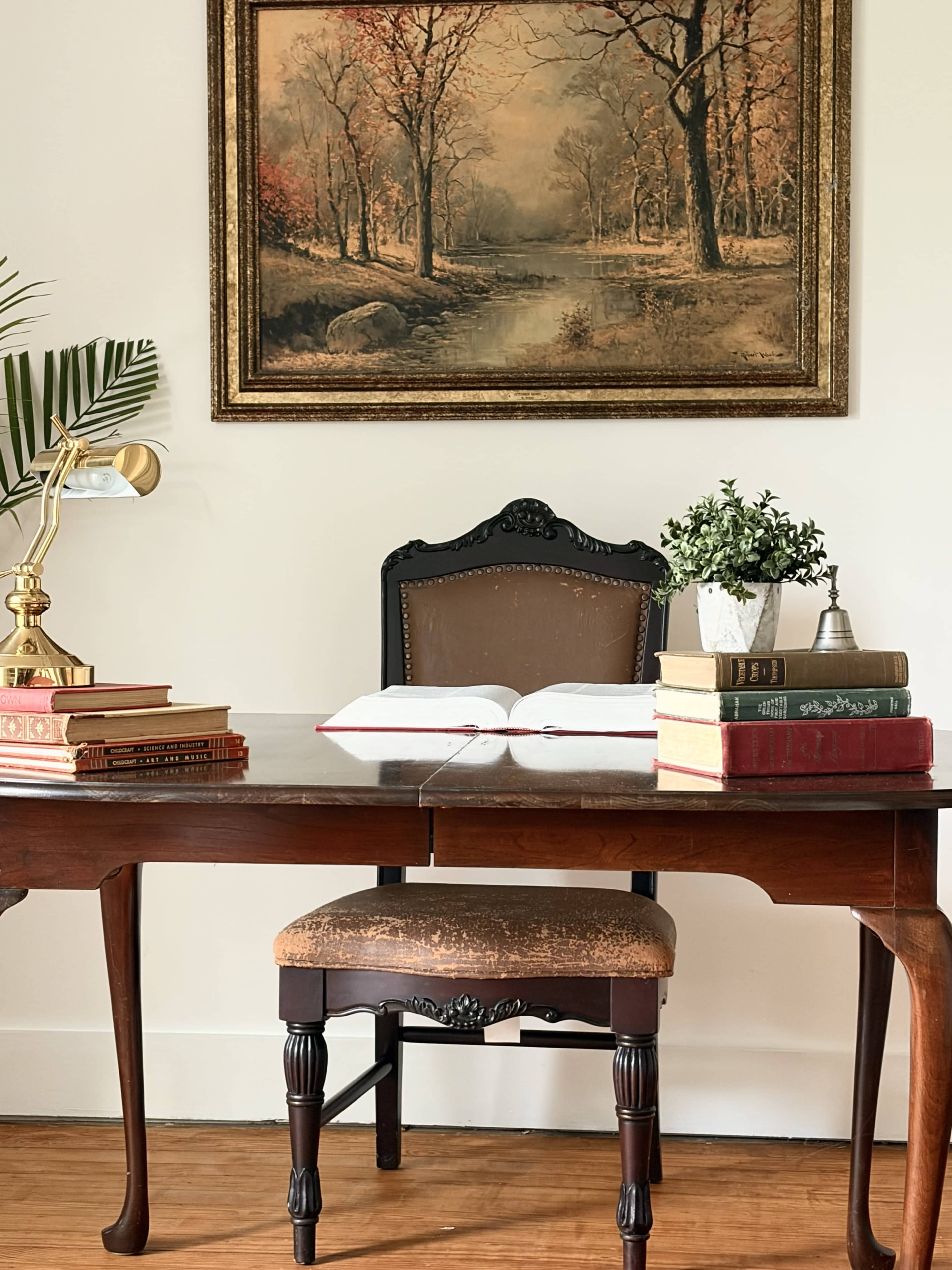 A wooden desk with an open book and stacked books sits in front of a chair, beneath a framed landscape painting, with a lamp and a small potted plant on the desk.