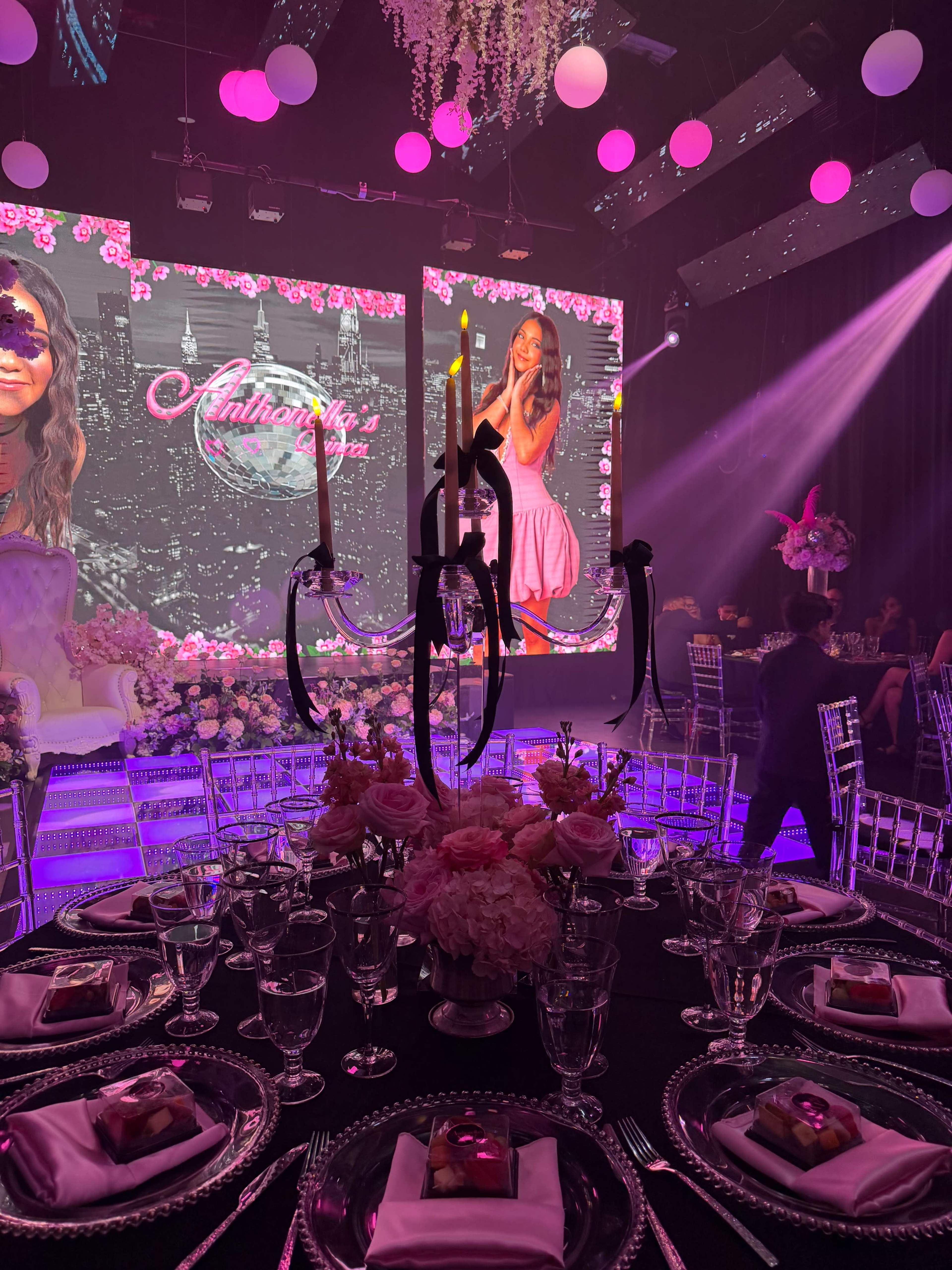 The image shows a lavish event setup featuring a round table adorned with elegant table settings, surrounded by chairs, with large screens displaying a floral-themed backdrop.
