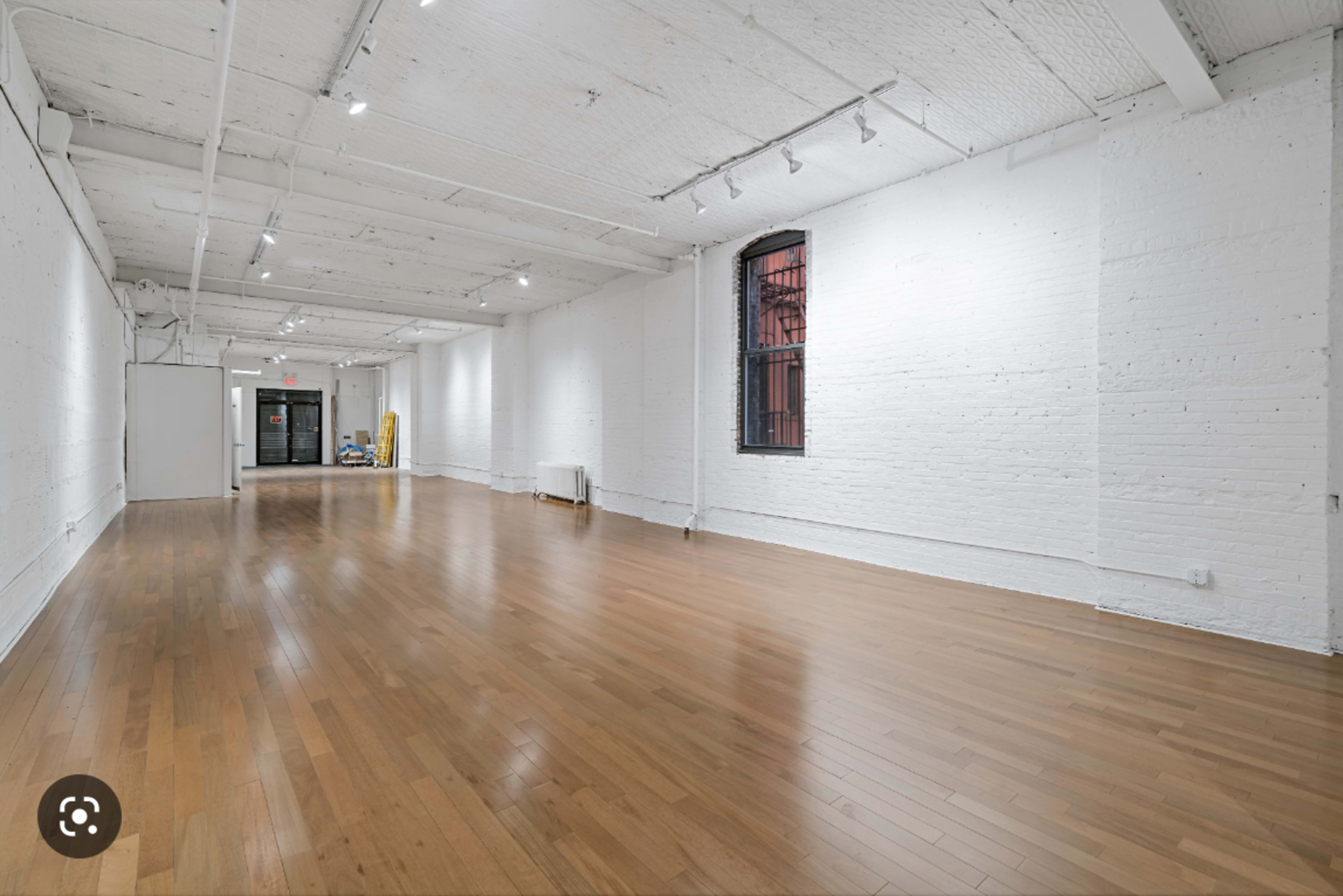 A spacious, empty room with wooden flooring and white brick walls, featuring large windows and track lighting.