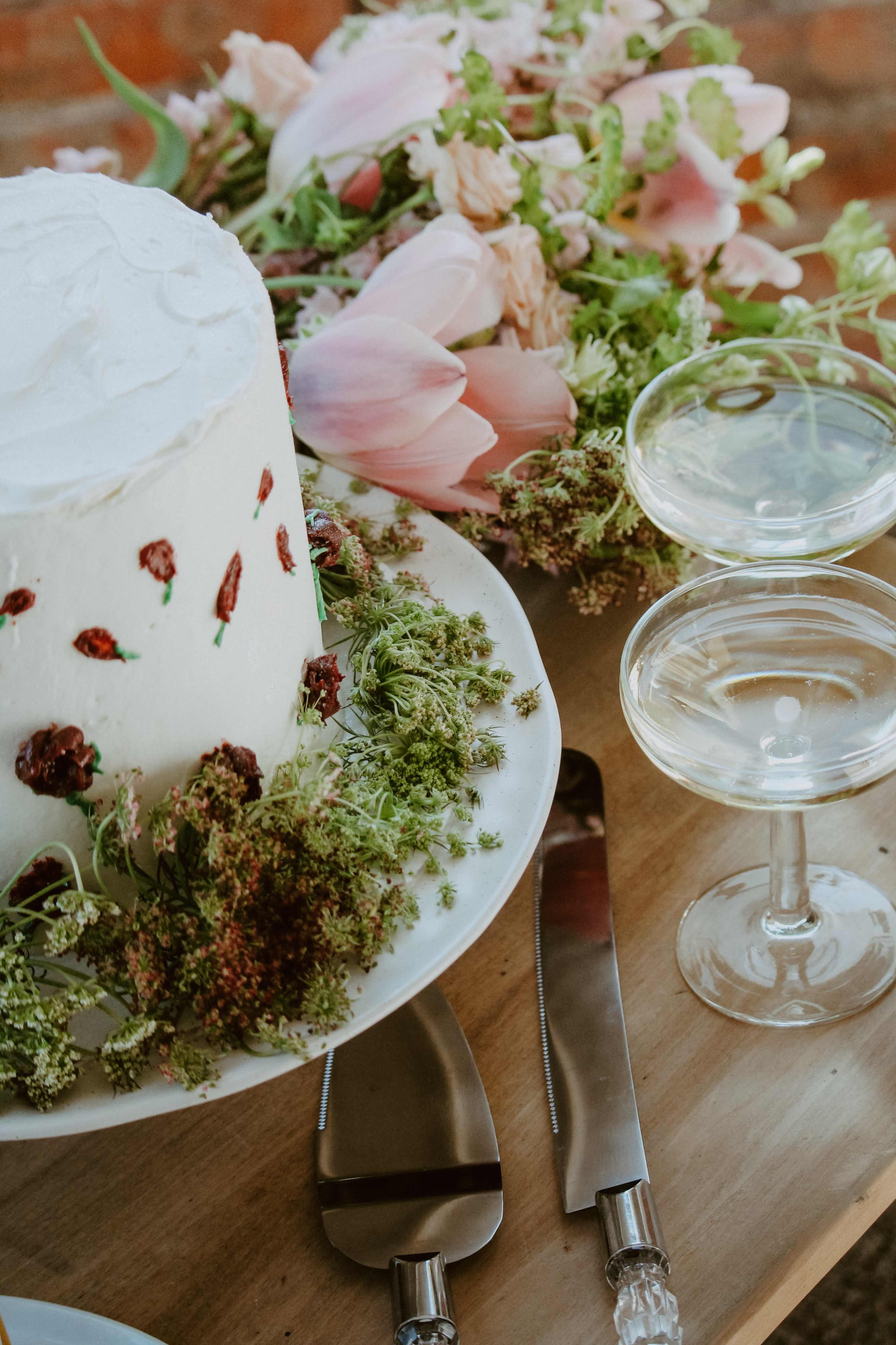 A white frosted cake adorned with dried flowers sits beside two champagne coupe glasses and a floral arrangement on a wooden table.