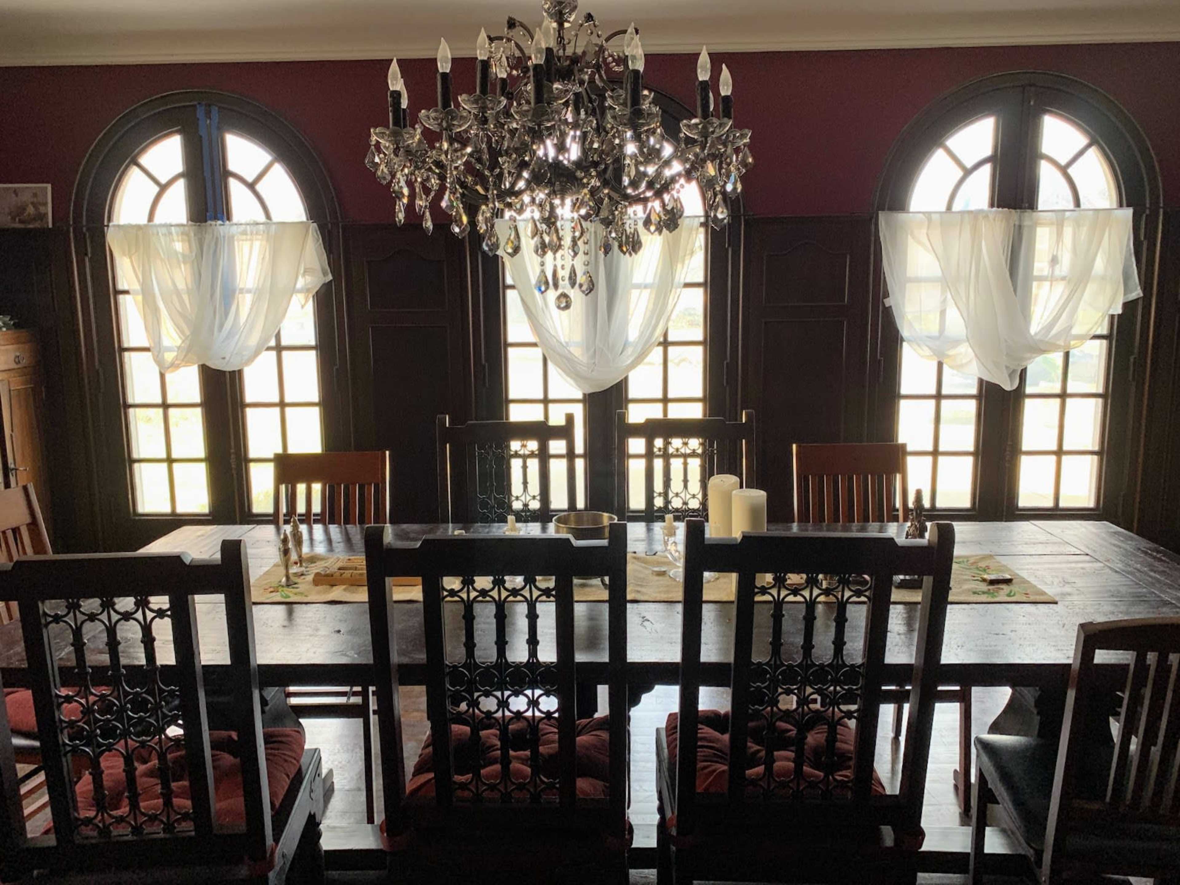 A large wooden dining table with eight chairs is set under a crystal chandelier, flanked by two arched windows dressed with sheer curtains.