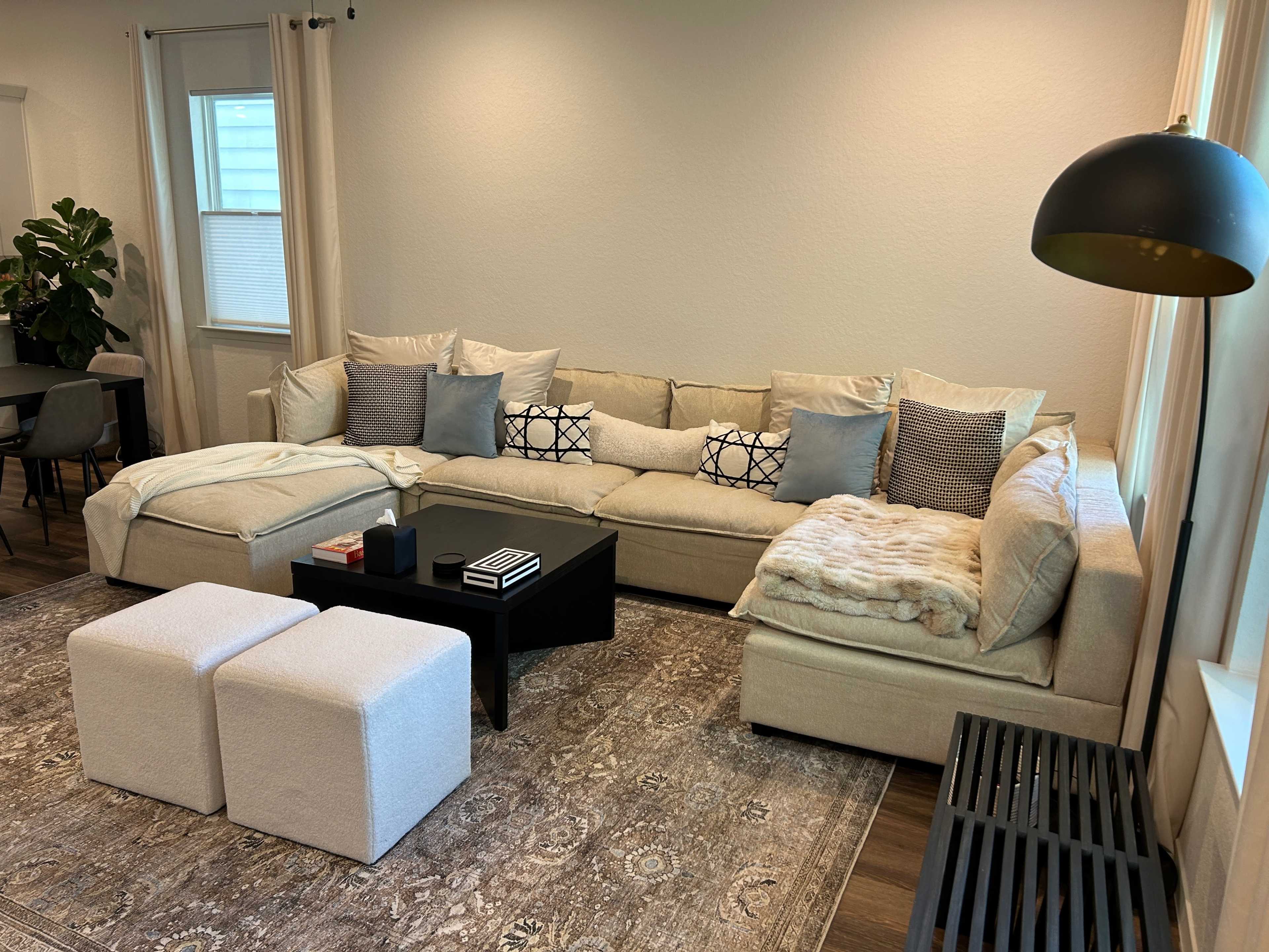 A spacious living room features a large beige sectional sofa with decorative pillows, a black coffee table, and two white ottomans on a patterned rug.