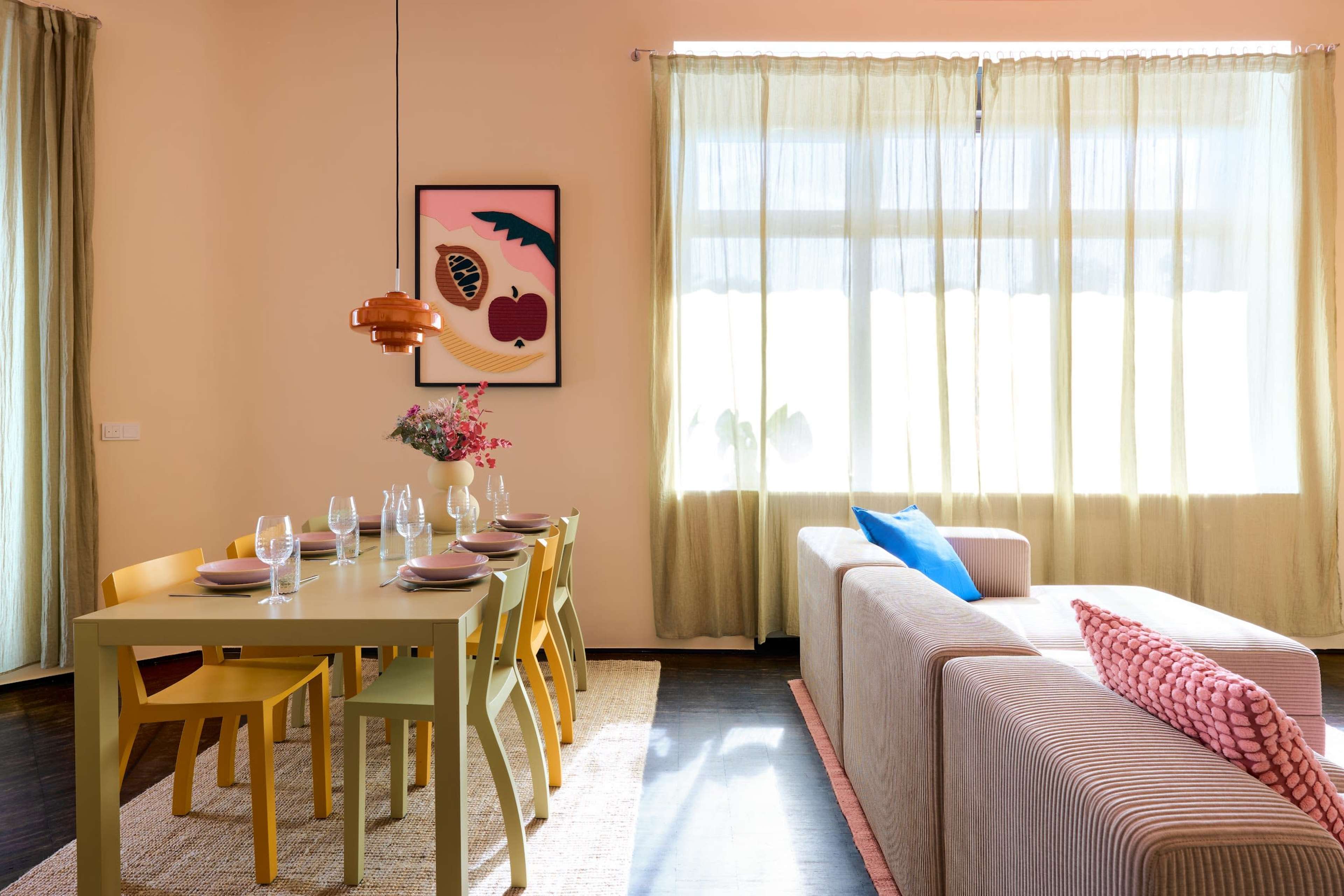 The image shows a modern dining area with a long table set for dinner, surrounded by colorful chairs, next to a light-filled living space featuring a sofa and decorative wall art.