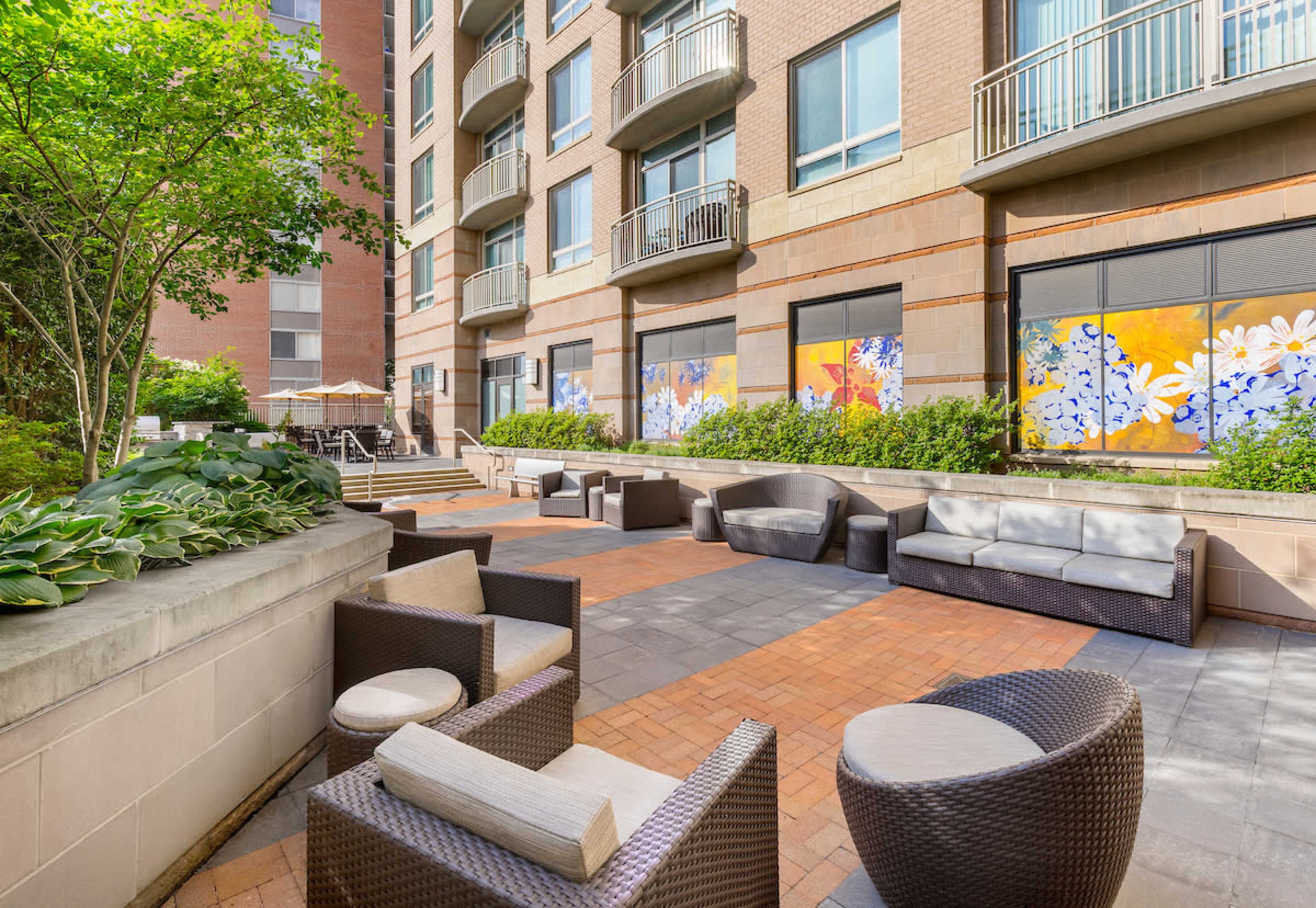 A patio area with modern furniture, including lounge chairs and tables, surrounded by greenery and a building with large windows.
