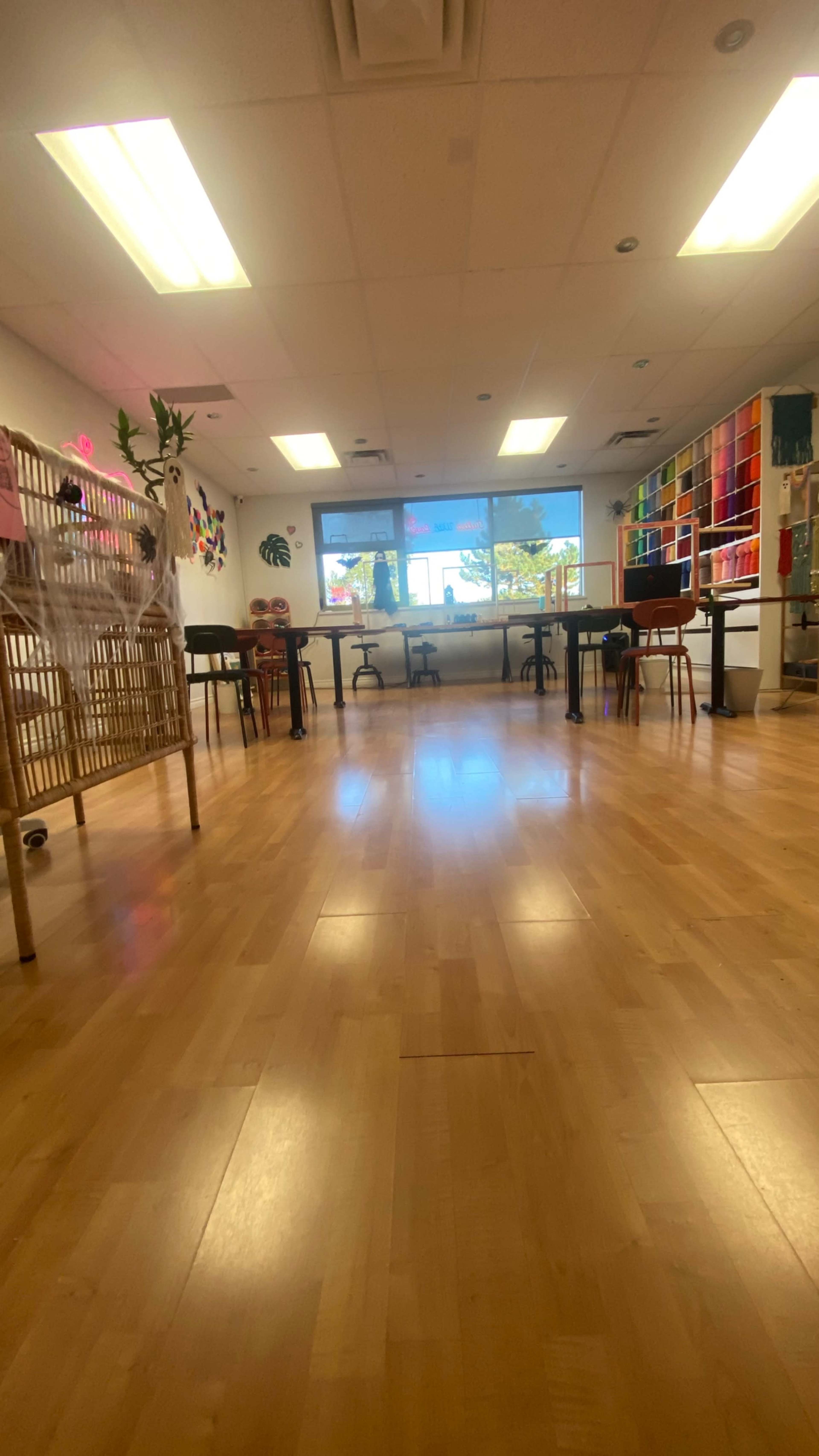 The image shows a bright, spacious room with wooden flooring, tables, and colorful shelves along the walls.