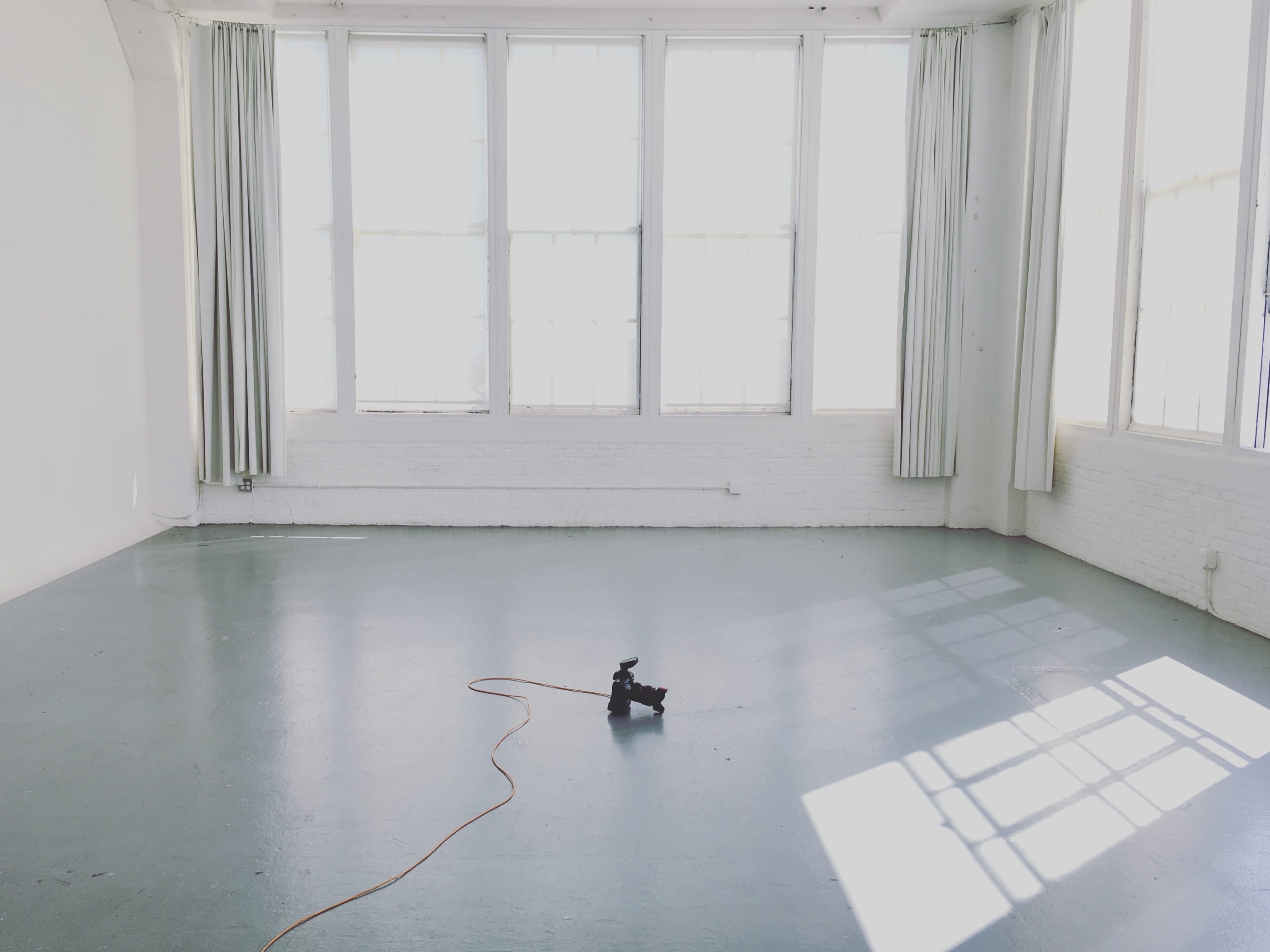 A spacious room with large windows, white walls, and a single black object on the floor connected by an orange extension cord.