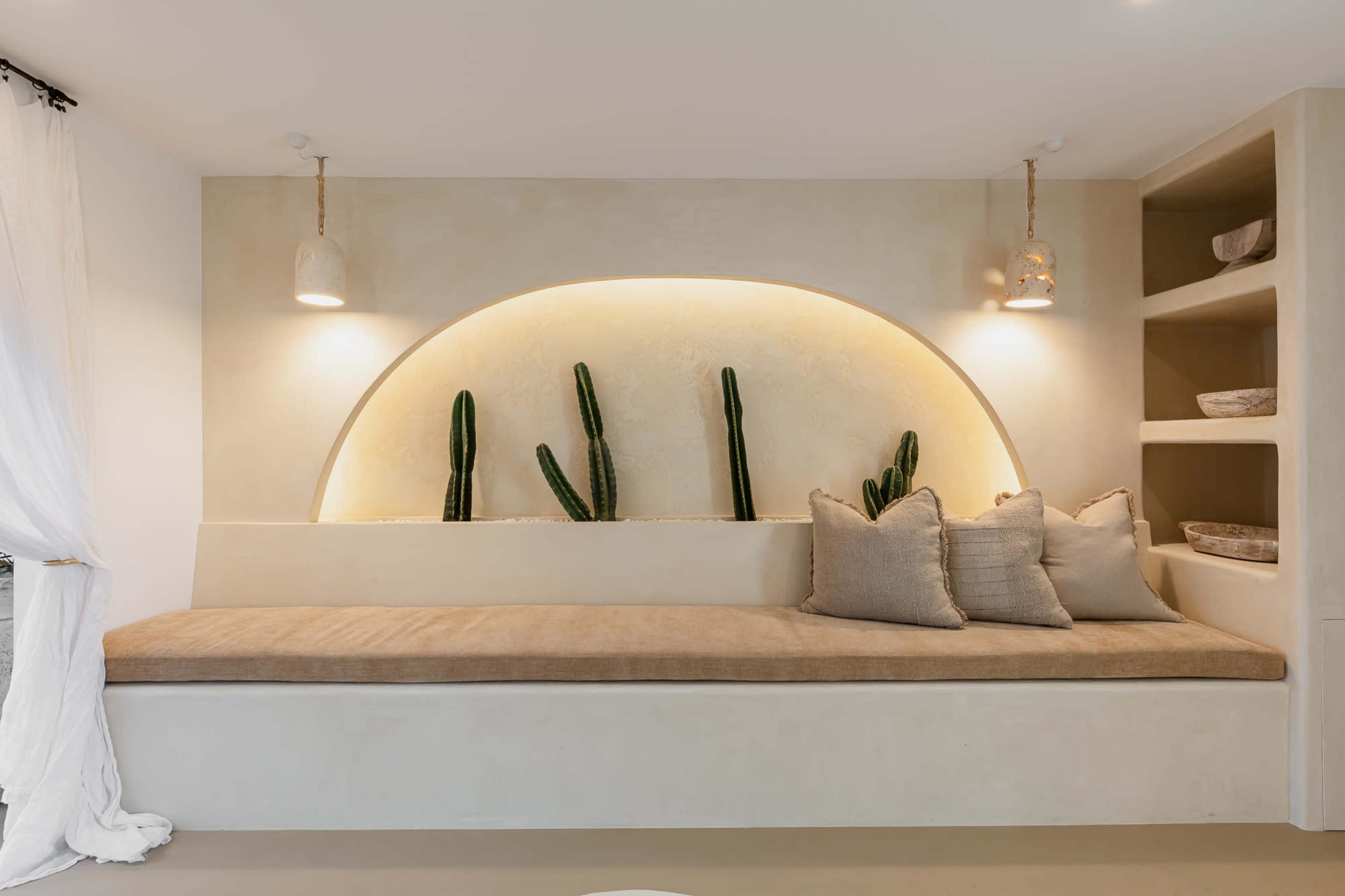 A minimalistic interior with a curved wall niche containing three upright cacti, a built-in bench with neutral cushions, and wall shelves holding decorative baskets.