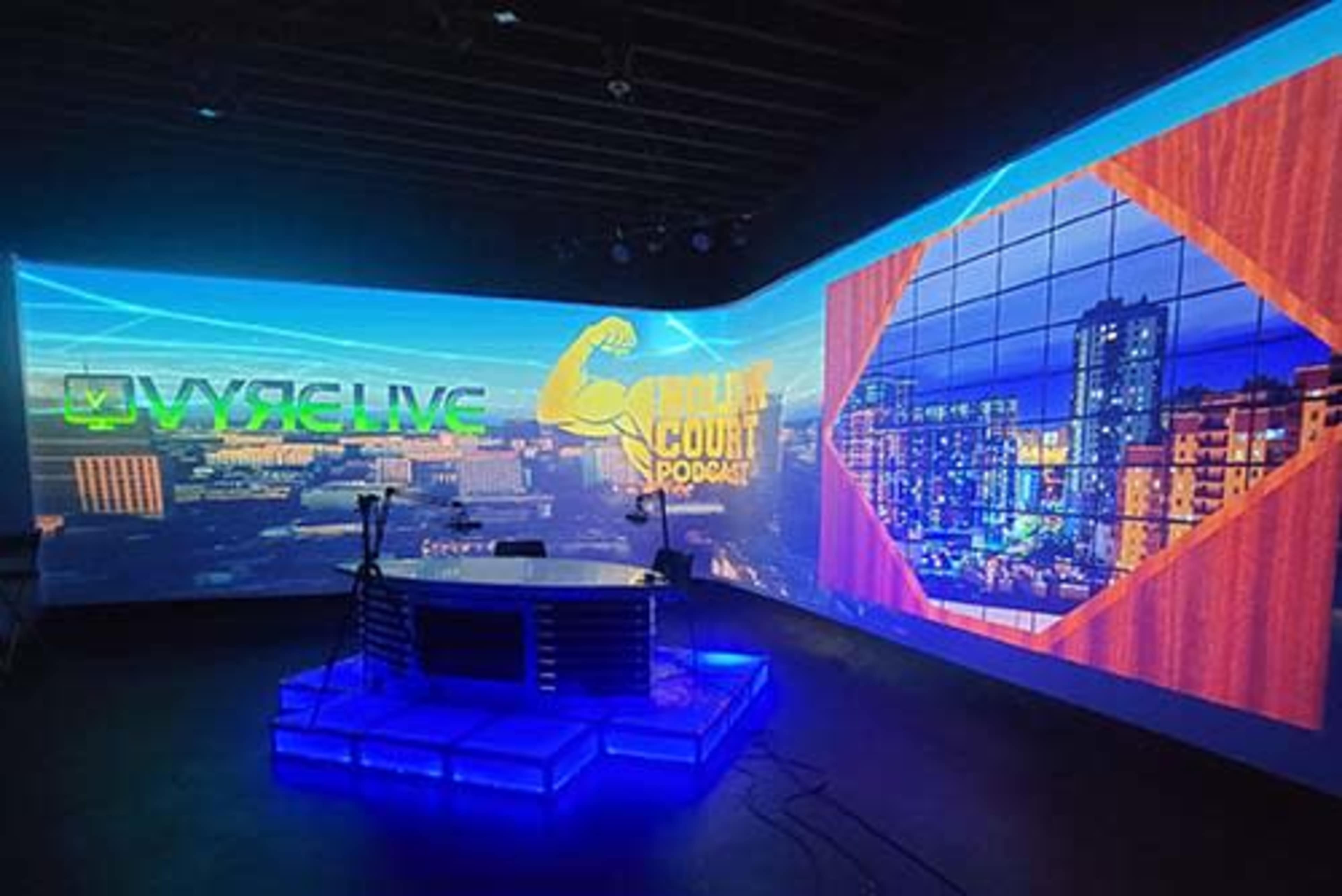 The image shows a modern studio setup featuring a broadcast table illuminated with blue light and large screens displaying vibrant graphics for "VYRELIVE" and "6th Court Podcast."
