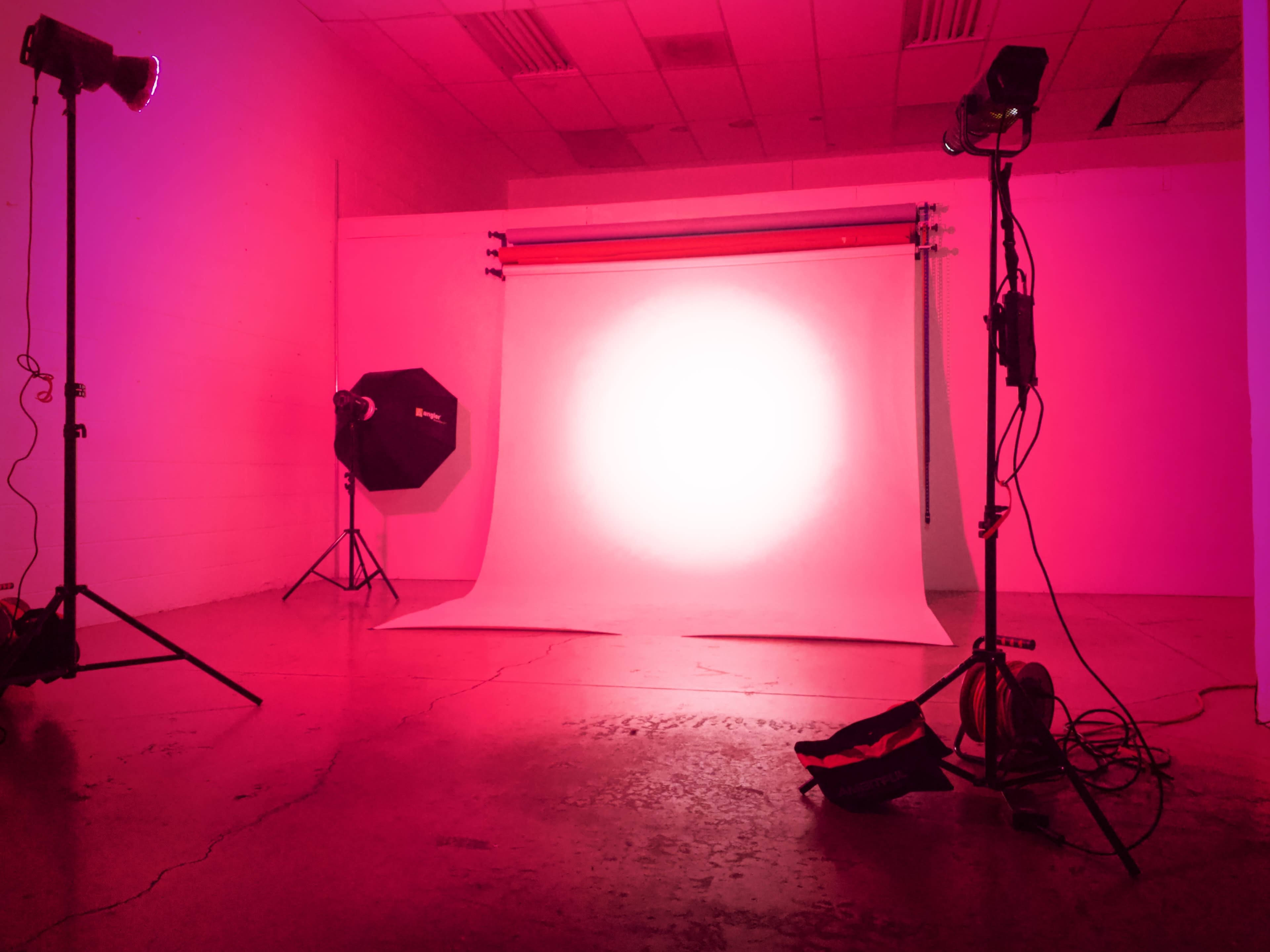 A photography studio with a pink backdrop, softbox, and lighting equipment positioned around a brightly lit area.