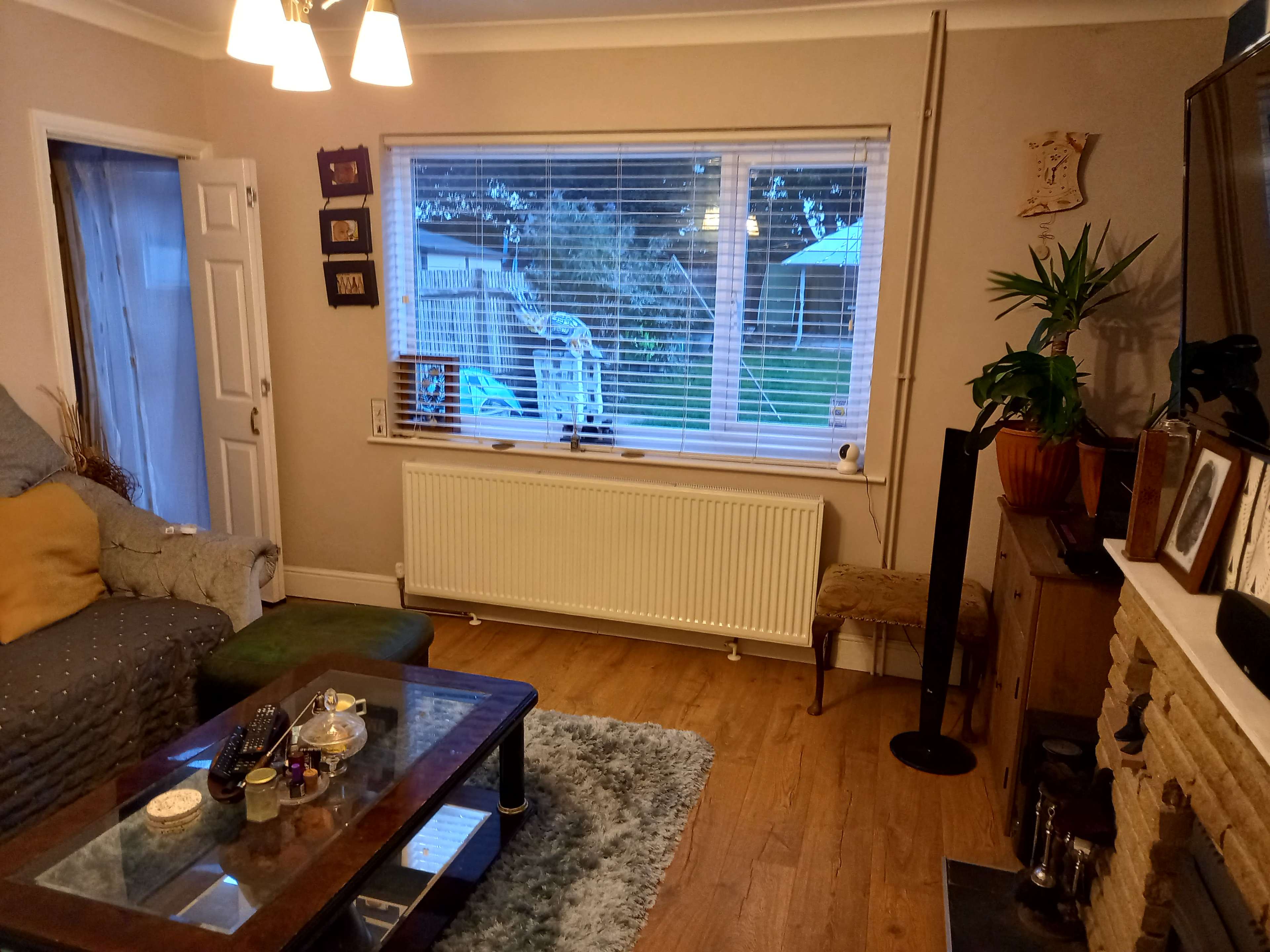 A cozy living room with a grey sofa, a coffee table displaying items, a large window with blinds, and a radiator underneath.