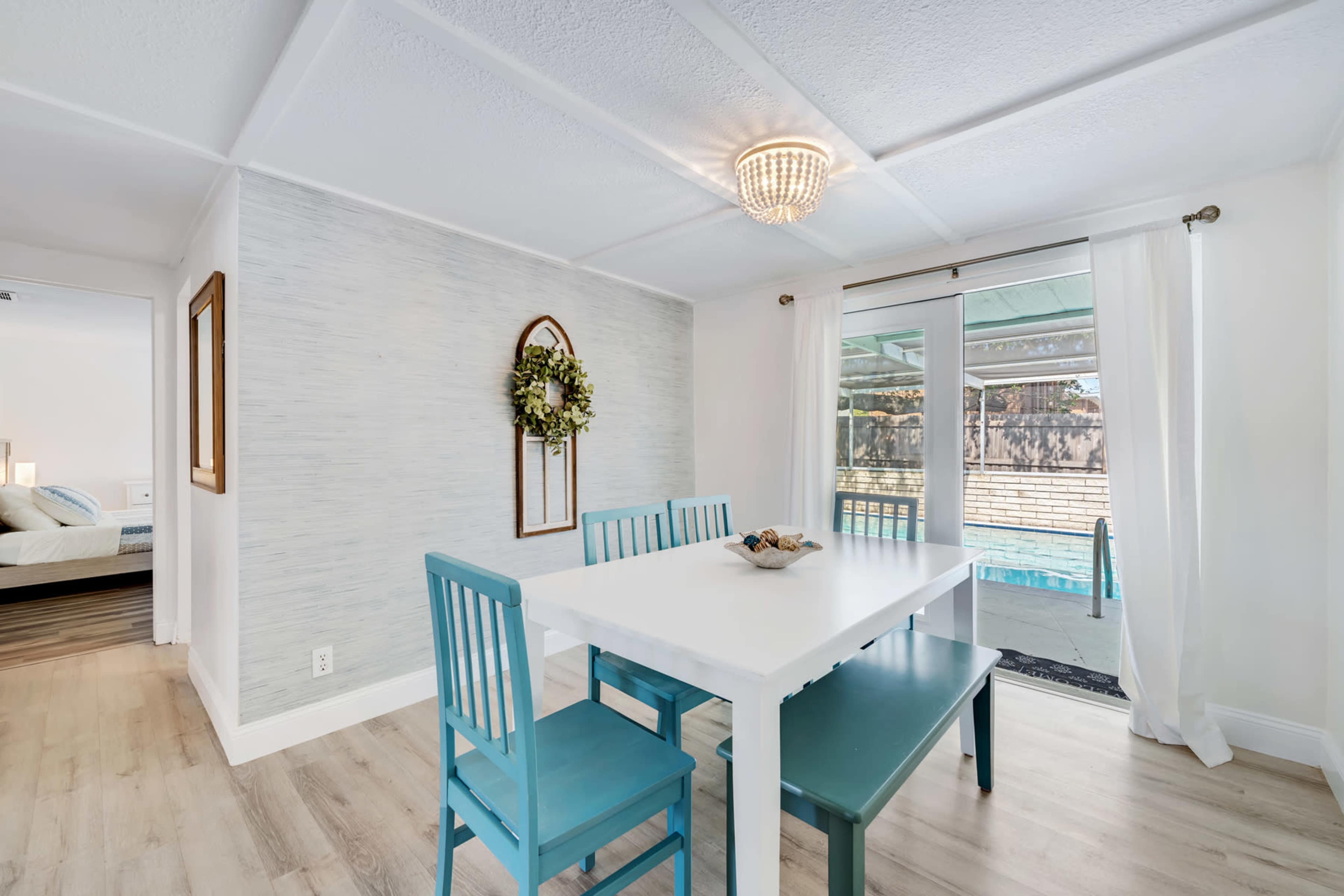A bright dining area features a white table surrounded by teal chairs, with a sliding glass door that opens to a pool view.