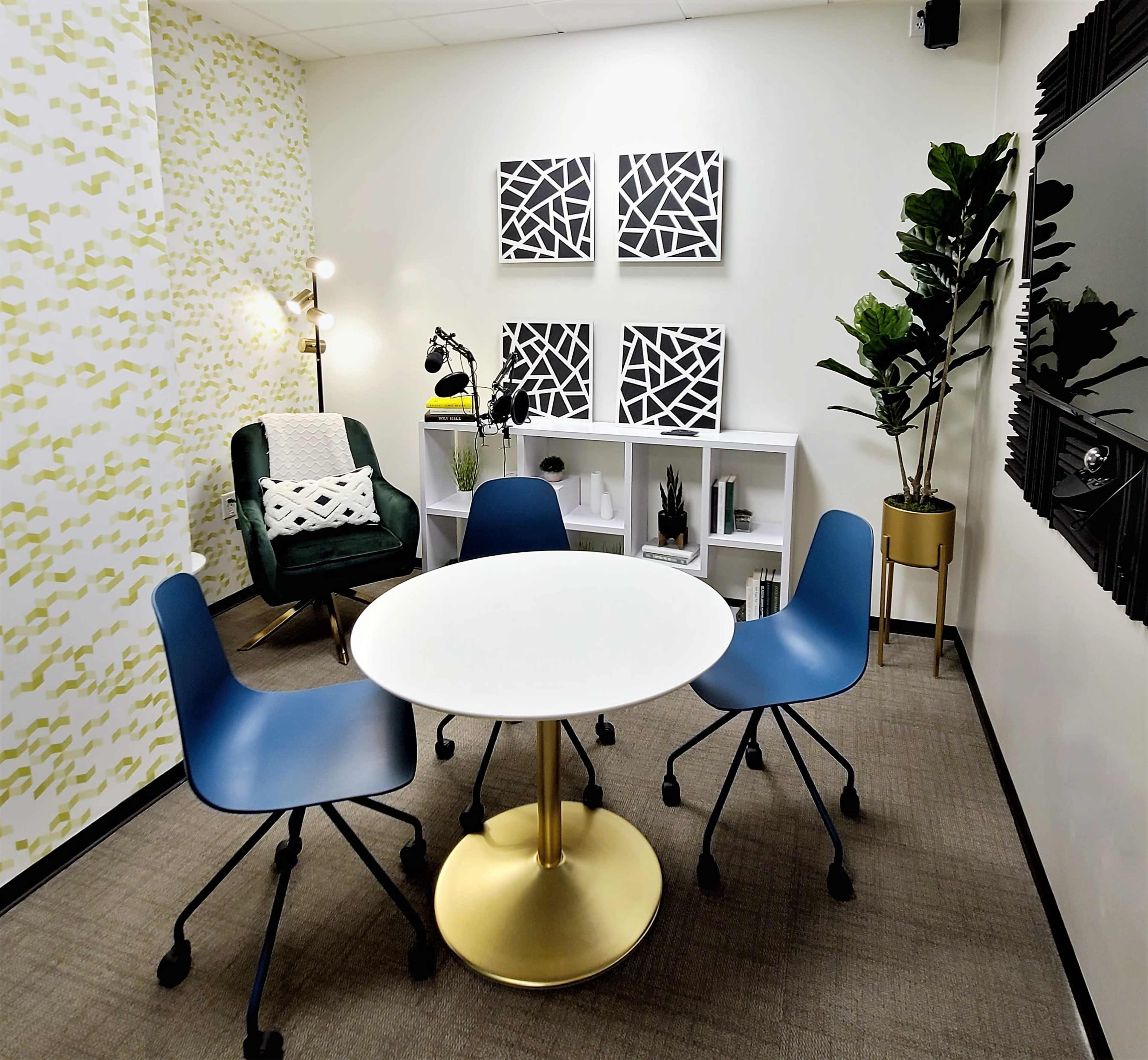 A small meeting room features a round white table surrounded by three blue chairs, with a green armchair, shelves, and black-and-white artwork on the walls.