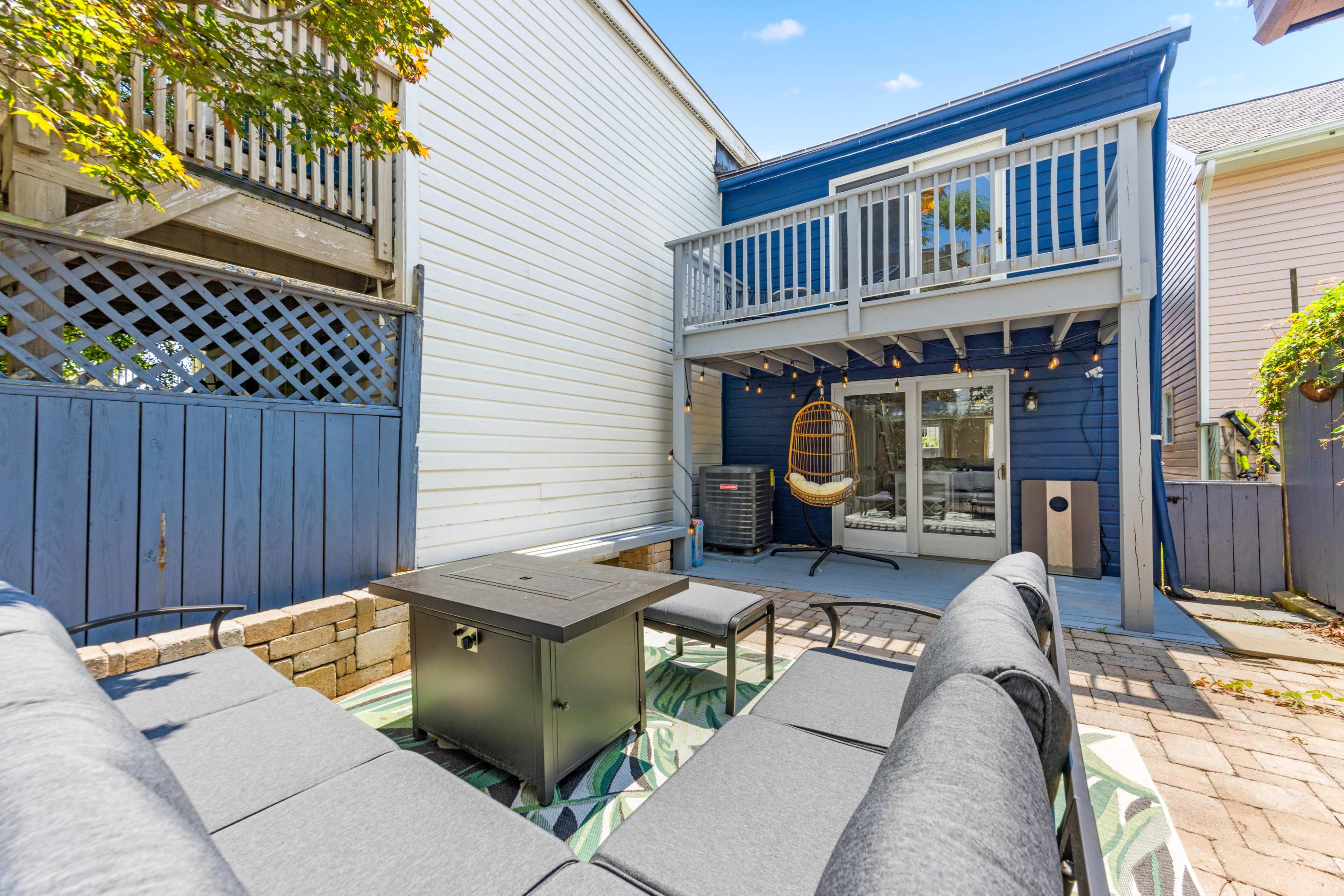The image shows a backyard patio area with gray outdoor furniture, a fire pit table, and a blue-and-white house in the background.