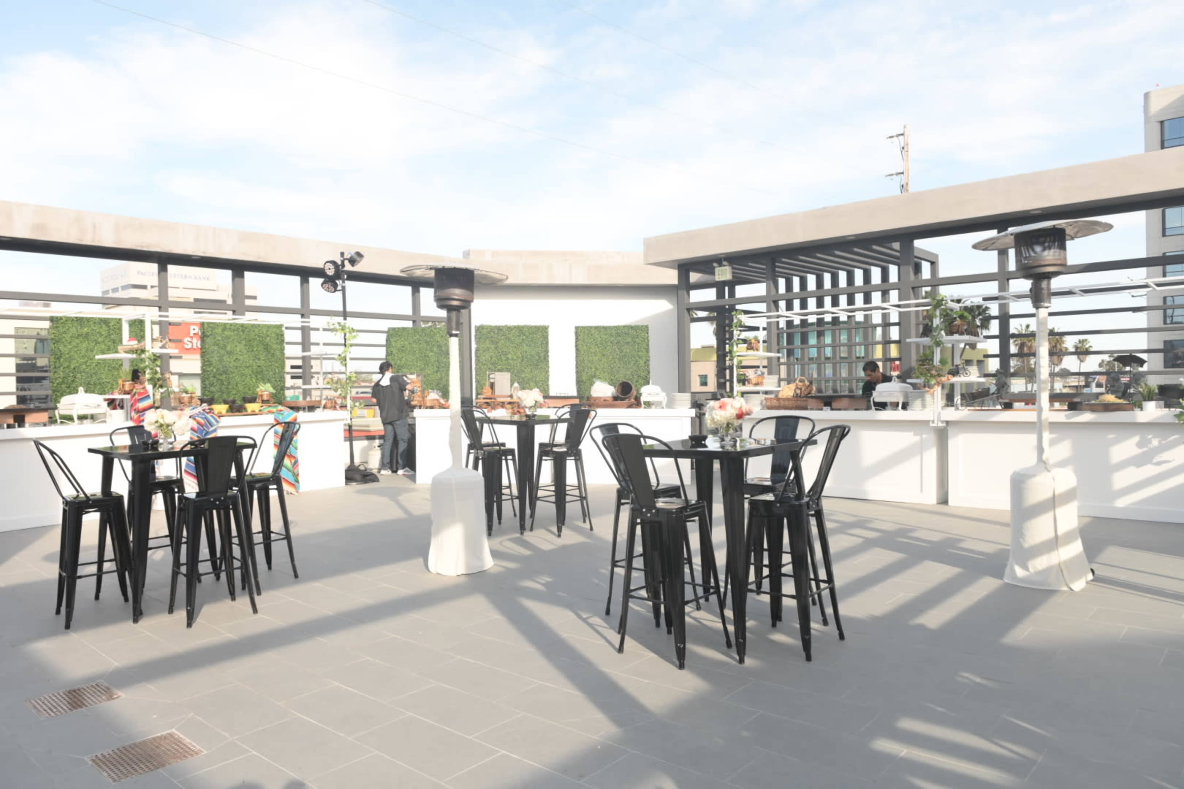A rooftop event space with black chairs and tables, surrounded by green foliage and equipped with heaters.