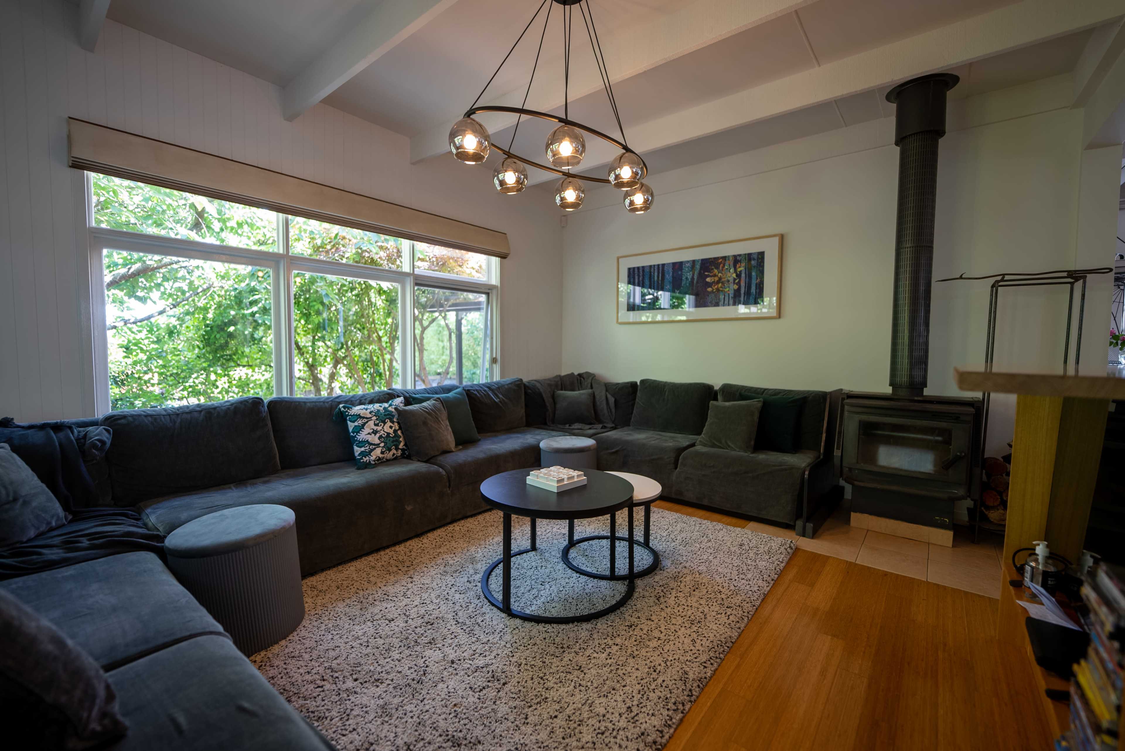 A spacious living room features a large sectional sofa, a circular coffee table, and a wood stove, with abundant natural light coming through large windows.