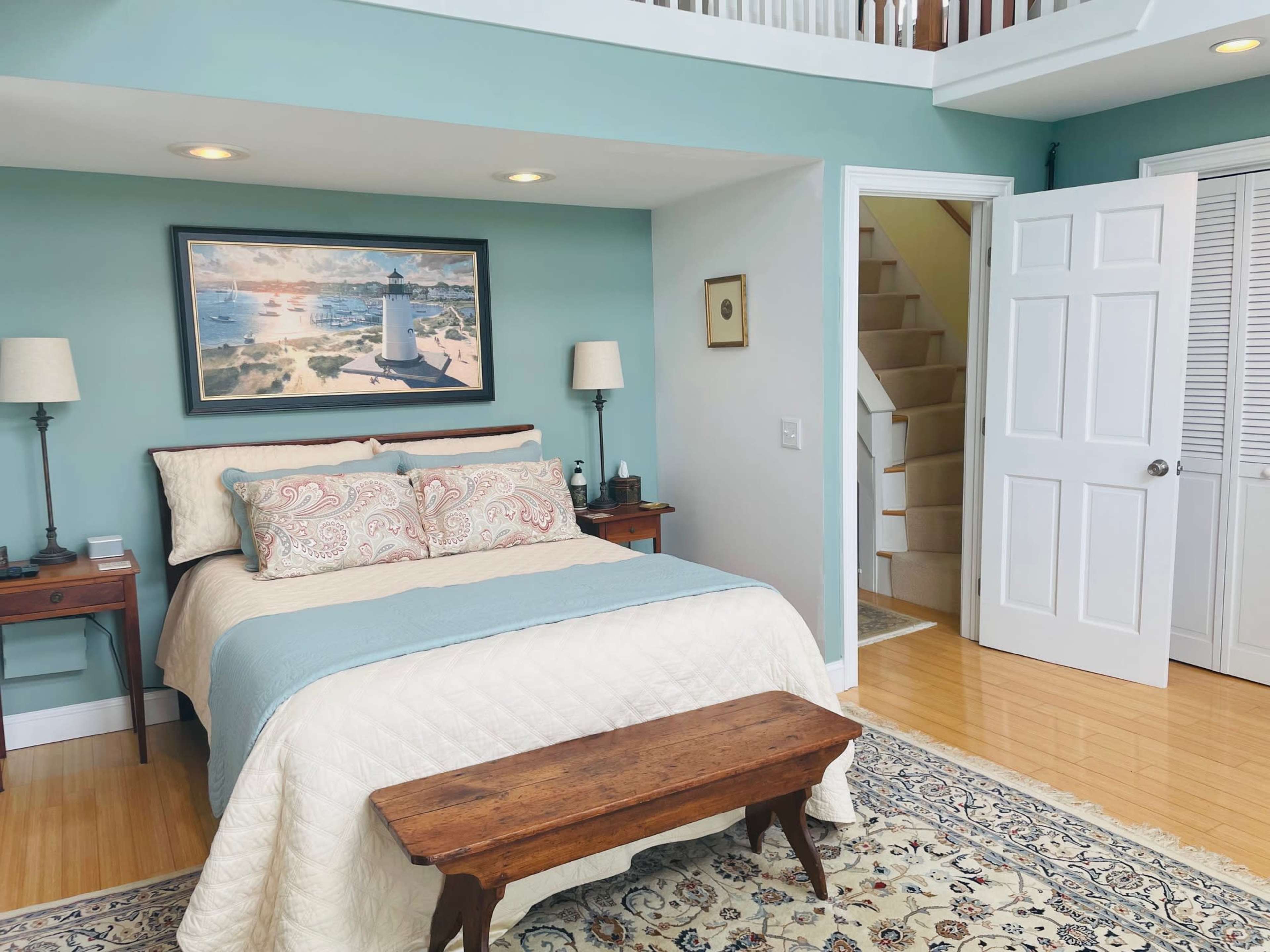 A cozy bedroom featuring a bed with decorative pillows, a wooden bench at the foot, a framed landscape painting on the wall, and an open door leading to a staircase.