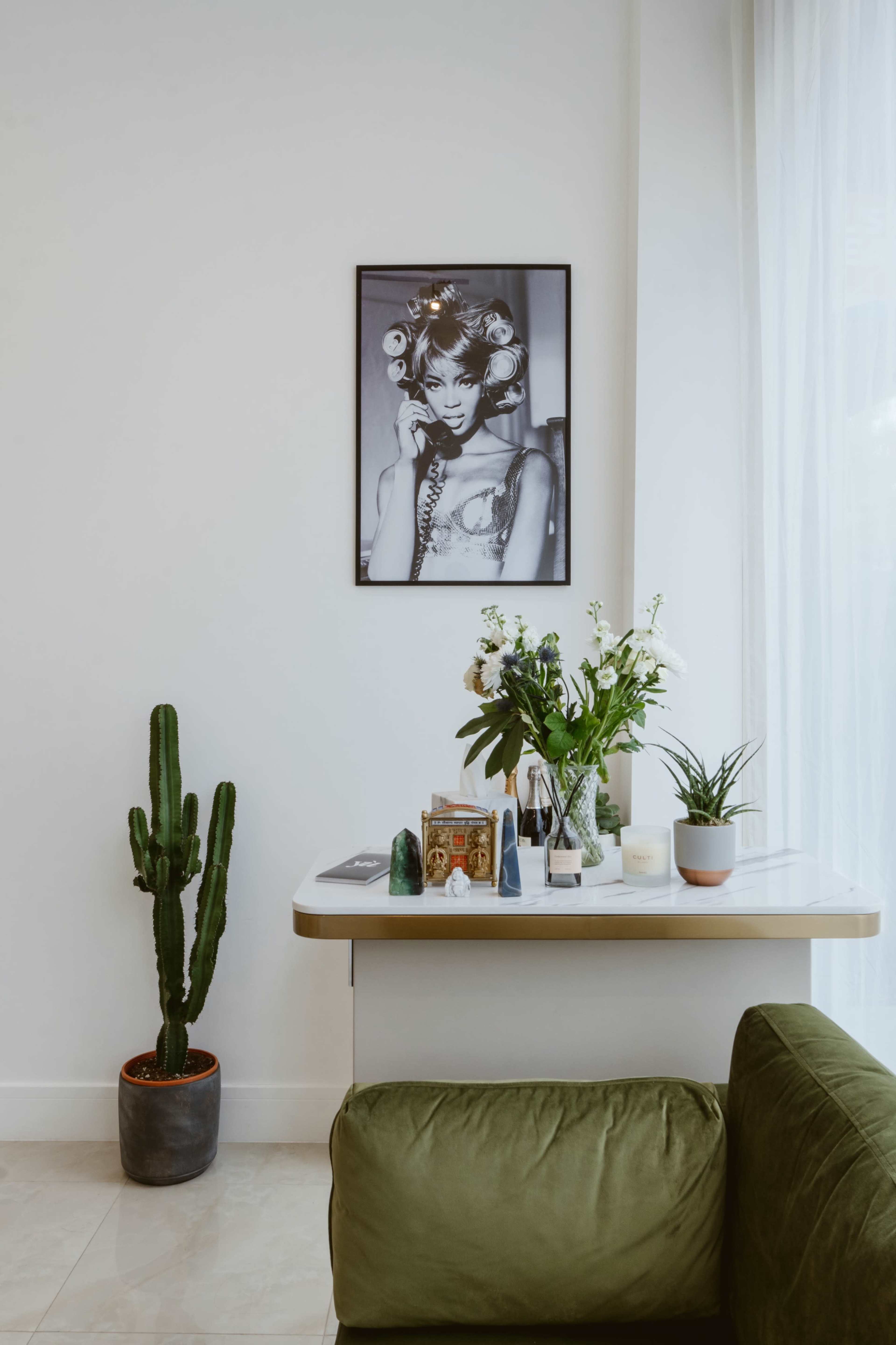 The image shows a minimalist interior with a large framed black-and-white portrait on the wall, a green couch, a potted cactus, and various decorative items on a table.